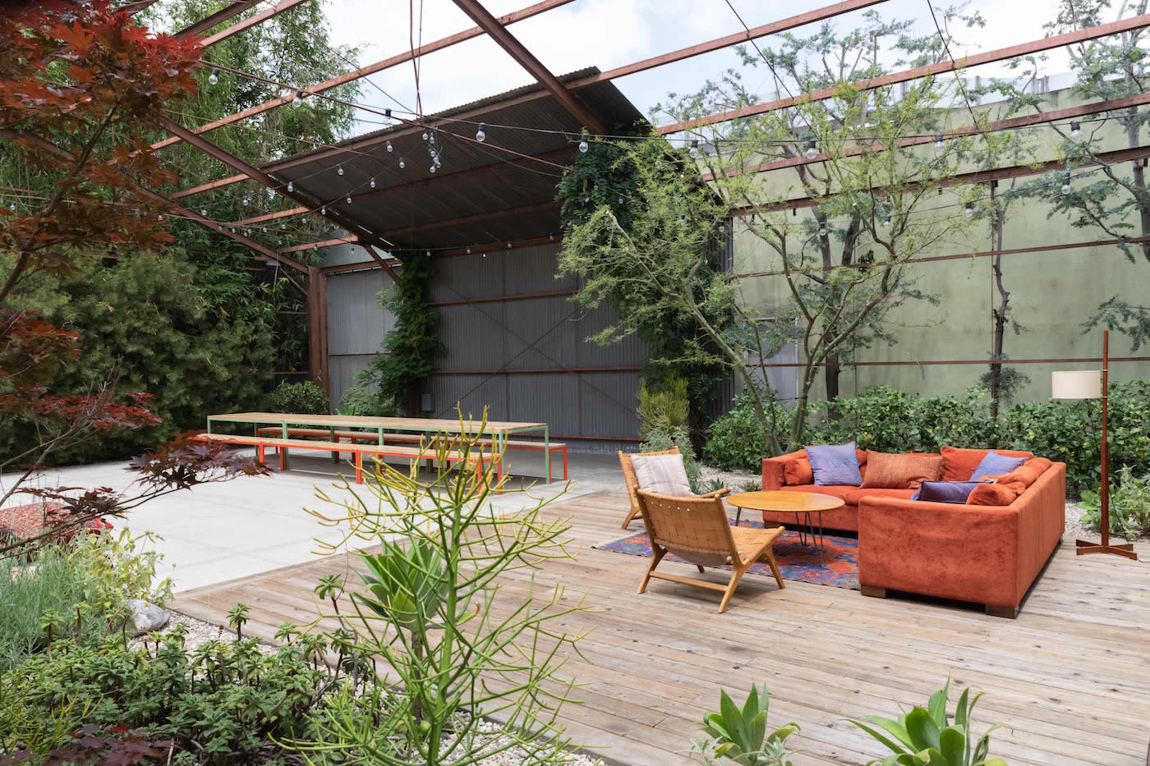 A modern outdoor seating area features a rusted metal framework surrounded by greenery, with a couch, two chairs, and a long table.