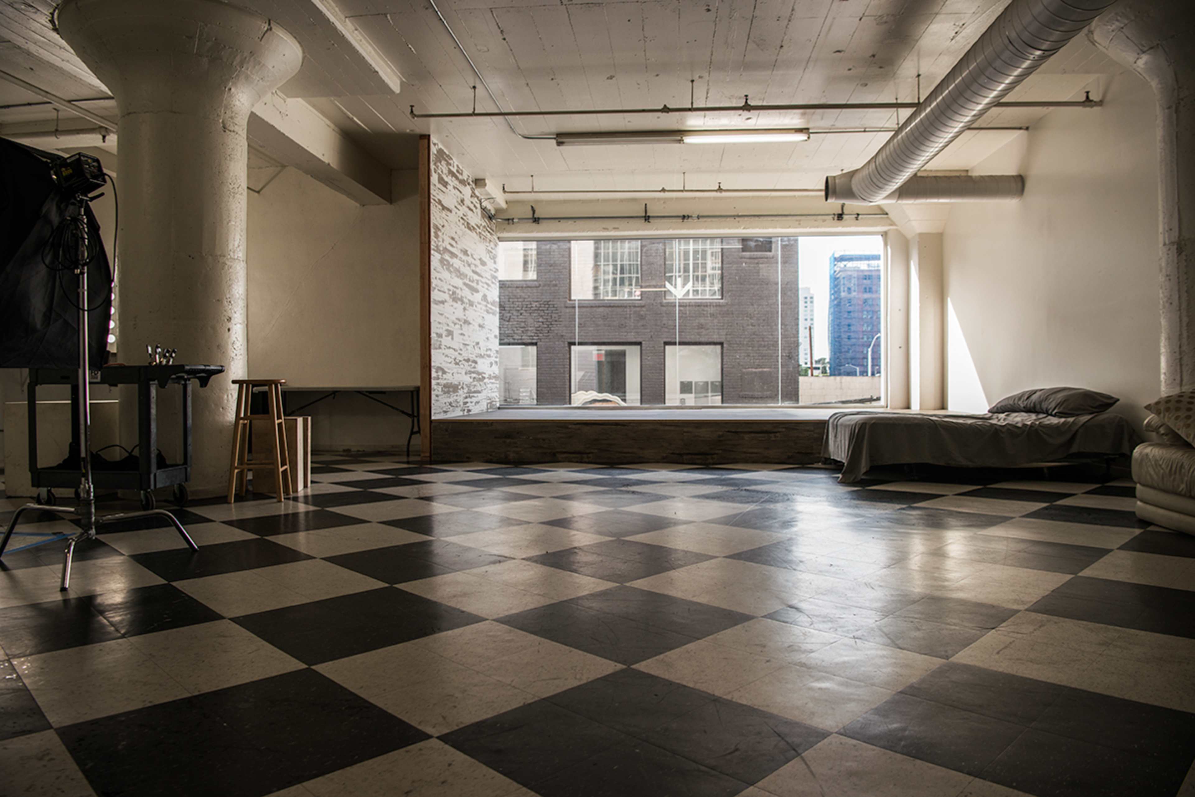 An empty, spacious room with a checkered floor, large windows, and minimal furnishings, including a bed and a stool.