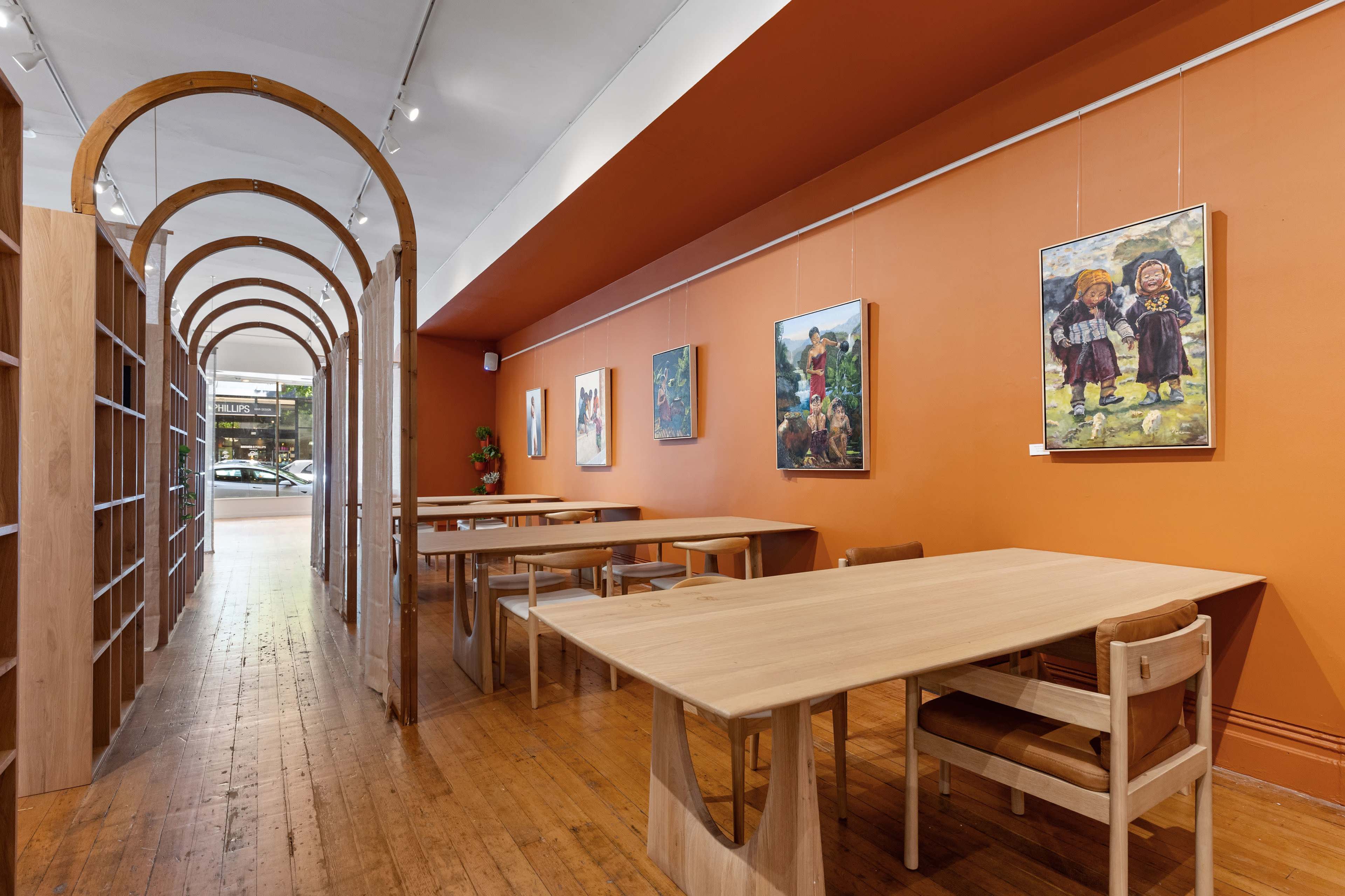 A well-lit interior of a café featuring wooden tables, an orange wall adorned with artwork, and an arched doorway leading to another area.