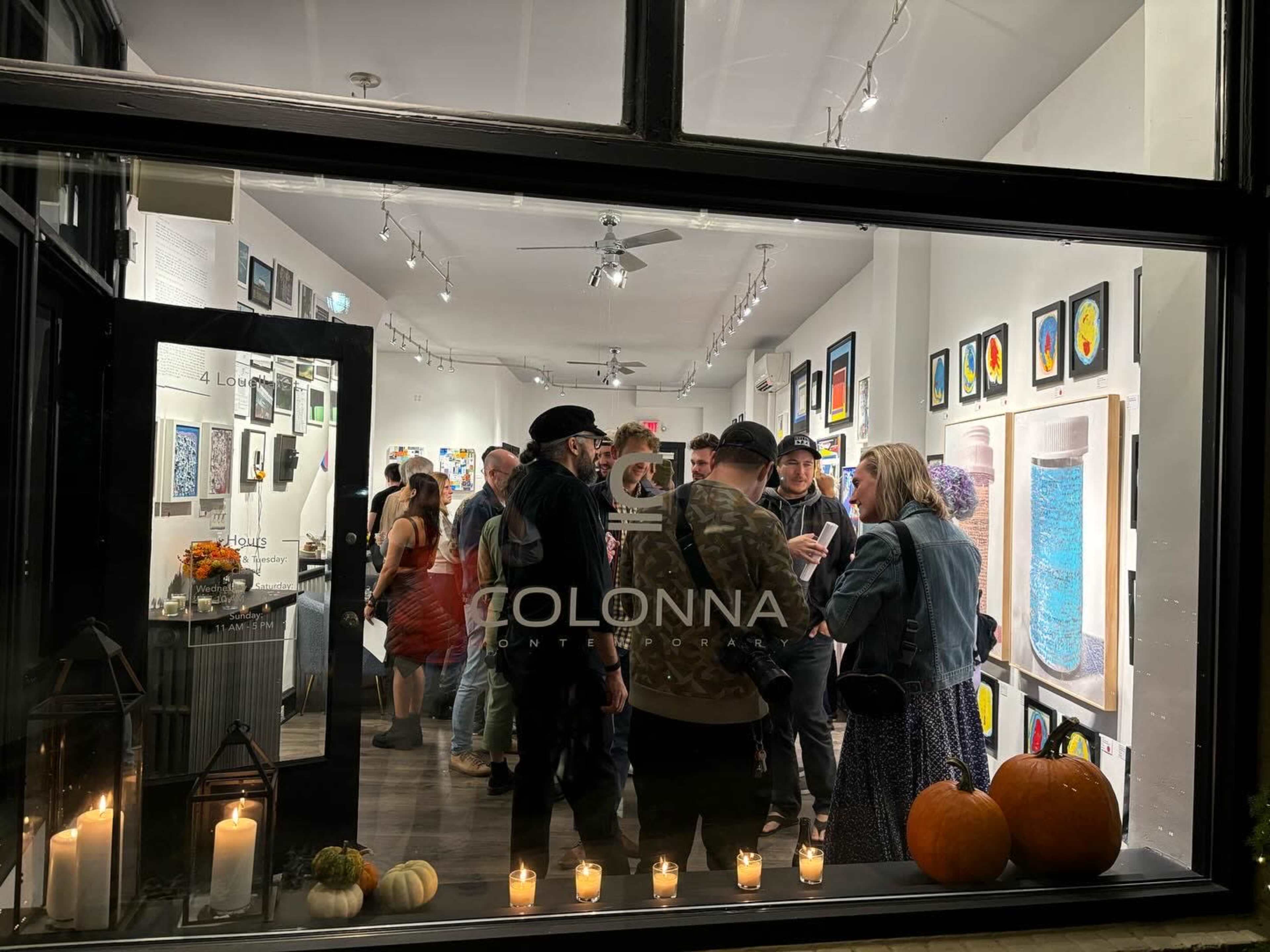 A group of people is gathered inside an art gallery, surrounded by colorful artworks displayed on the walls, with pumpkins and candles placed at the window.