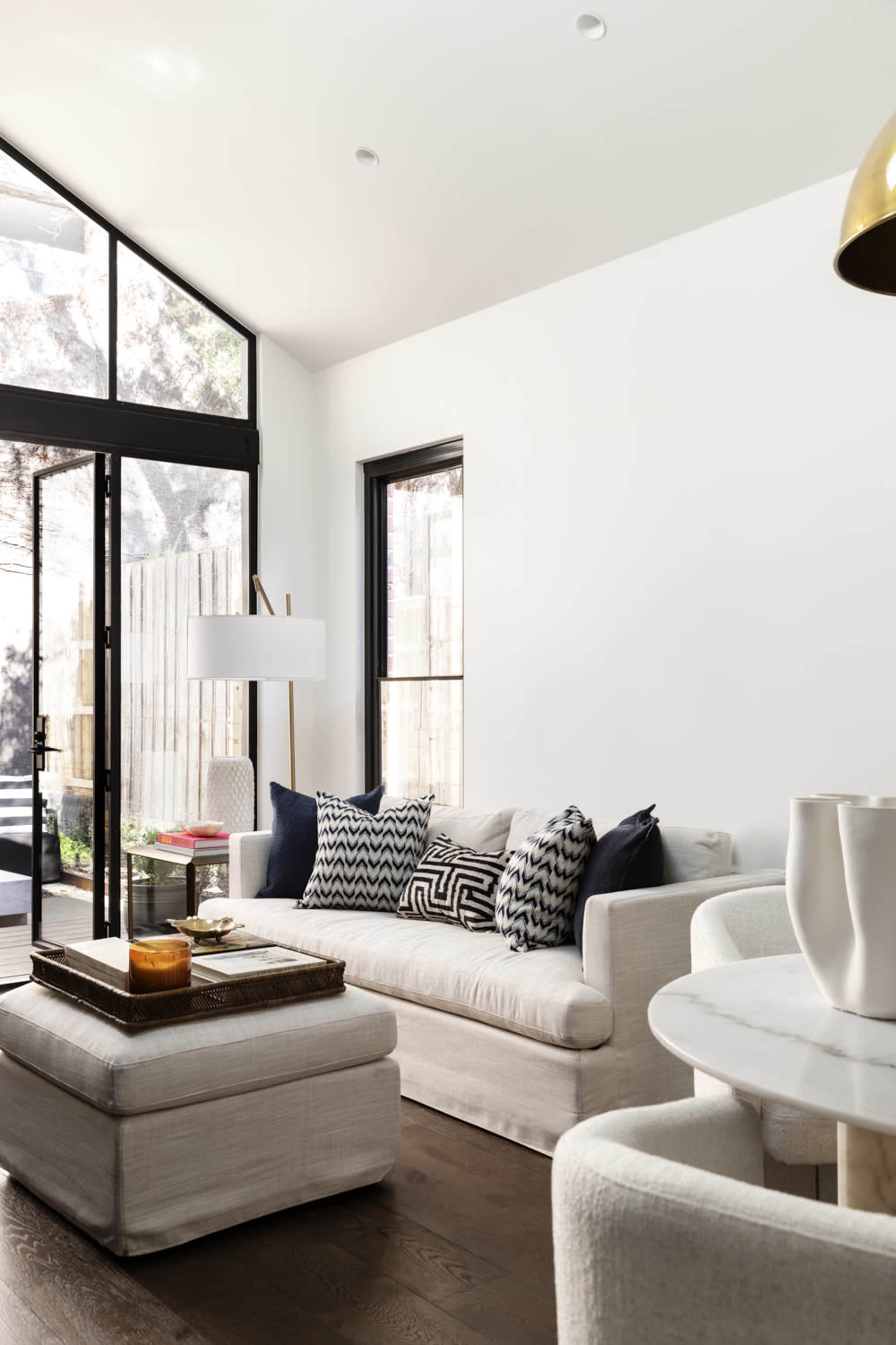 A modern living room with a light-colored sofa adorned with patterned cushions, a glass door leading to an outdoor space, and a round dining table with white vases.