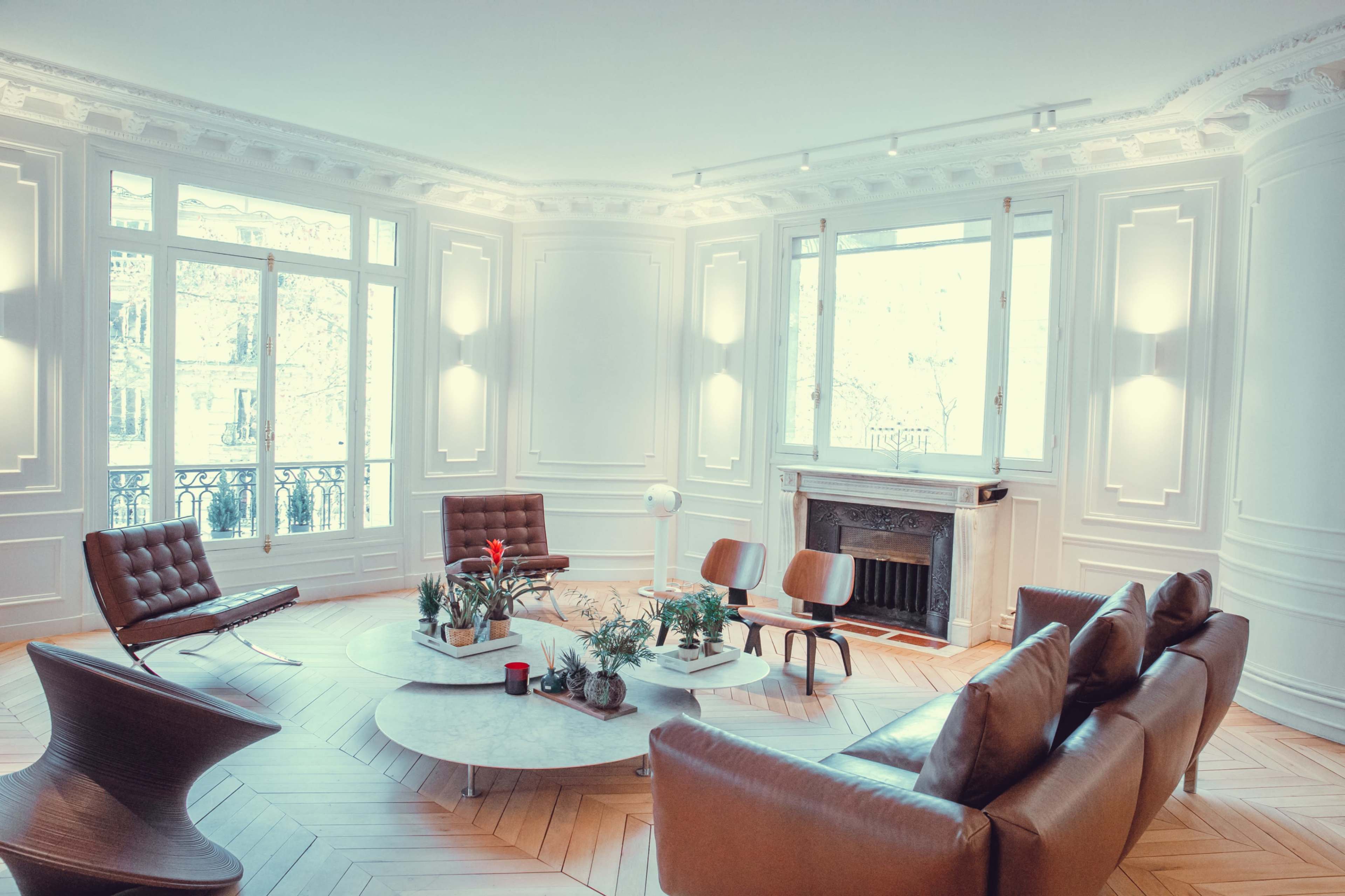 The image depicts a bright, modern living room featuring a symmetrical arrangement of furniture, including leather chairs and a circular coffee table, with large windows allowing natural light to enter.