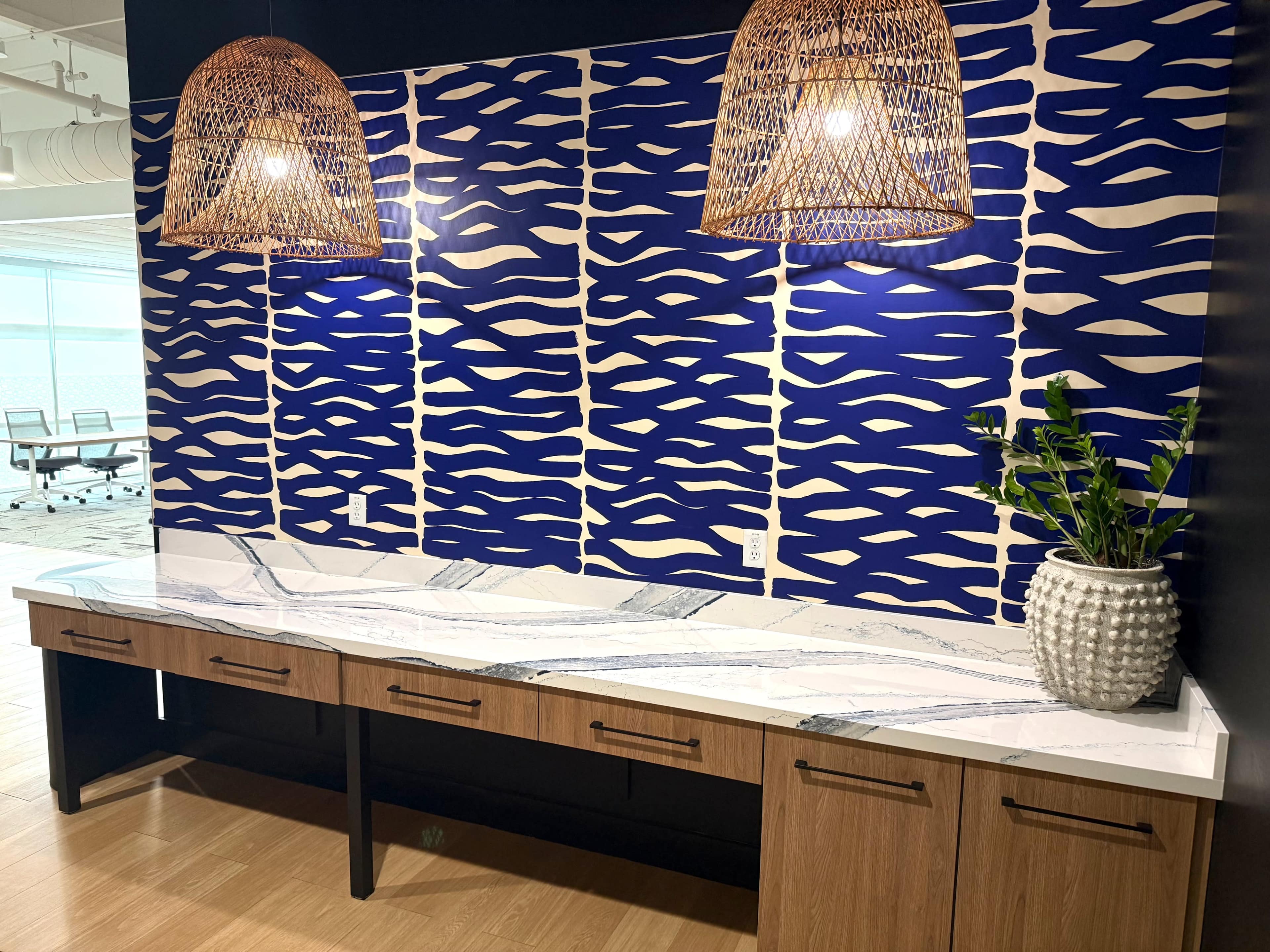 The image shows a modern workspace featuring a wooden desk with a marble countertop, complemented by a patterned blue wall and two woven pendant lights hanging above.