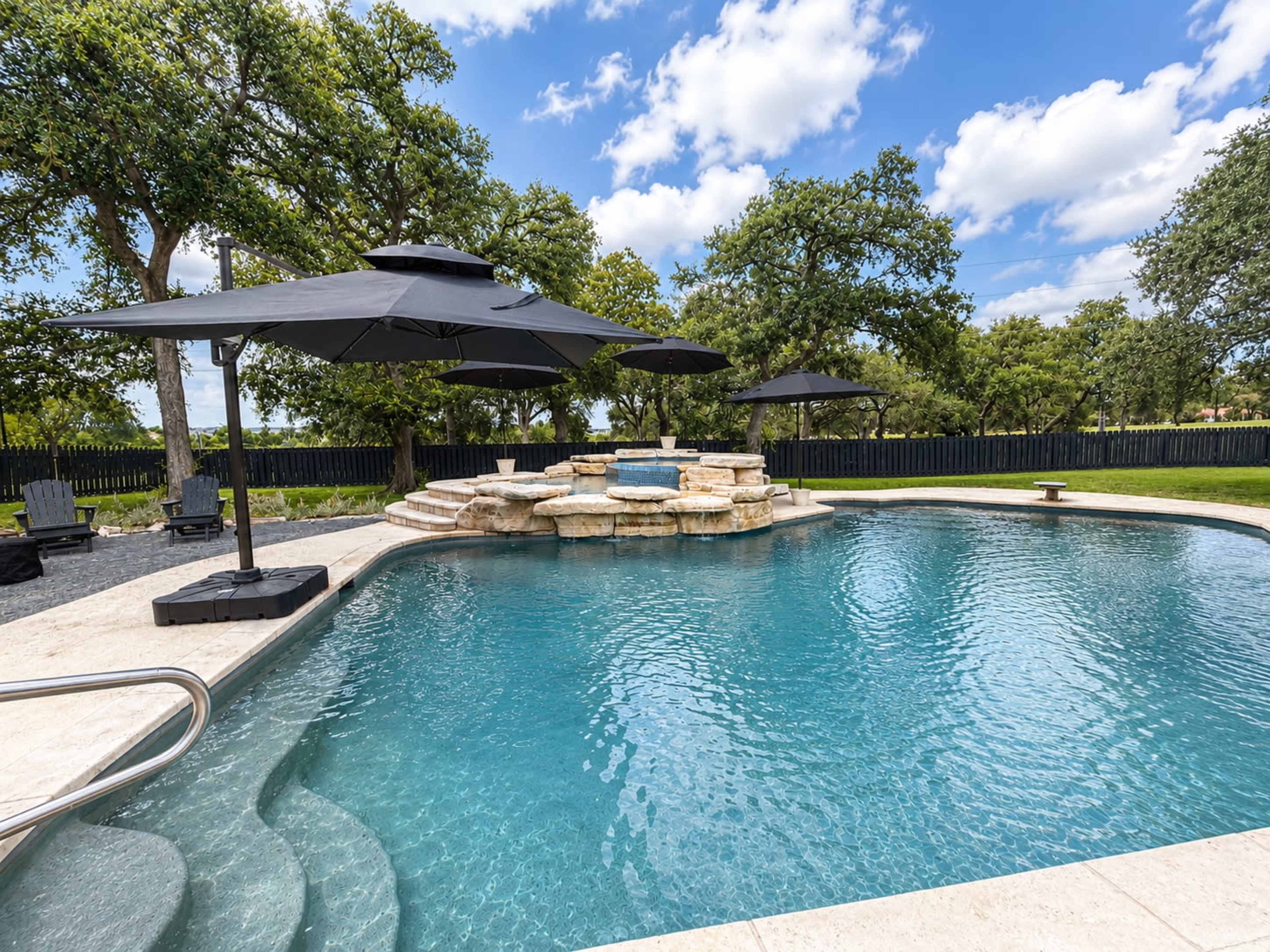 The image shows a swimming pool featuring a rocky spa area, surrounded by black umbrellas and lush greenery under a blue sky with fluffy clouds.