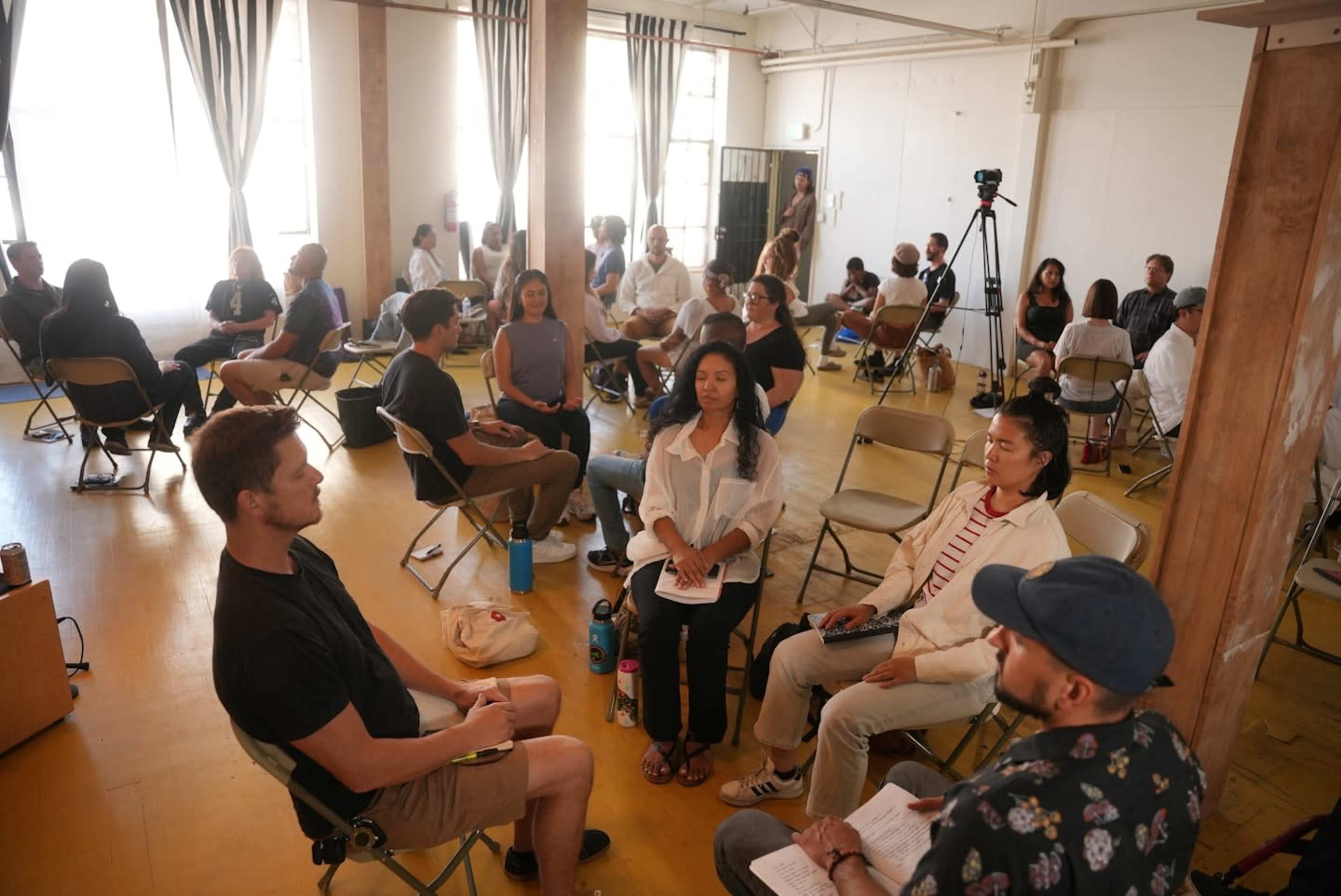 A group of people is seated in a large room, engaged in a discussion or workshop, with chairs arranged in a circle.