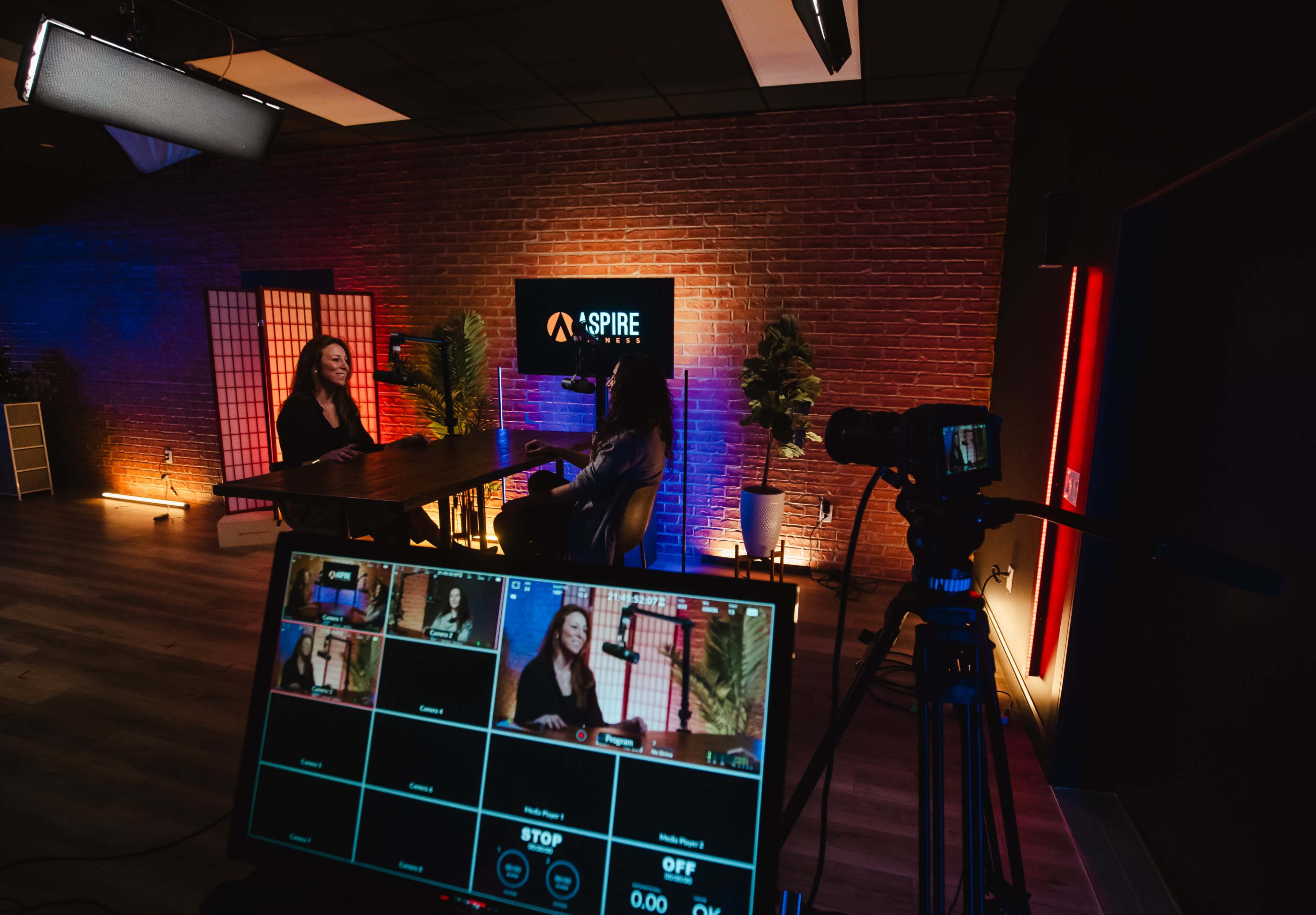 A camera setup captures a conversation between two individuals in a well-lit studio with a brick wall and modern decor.