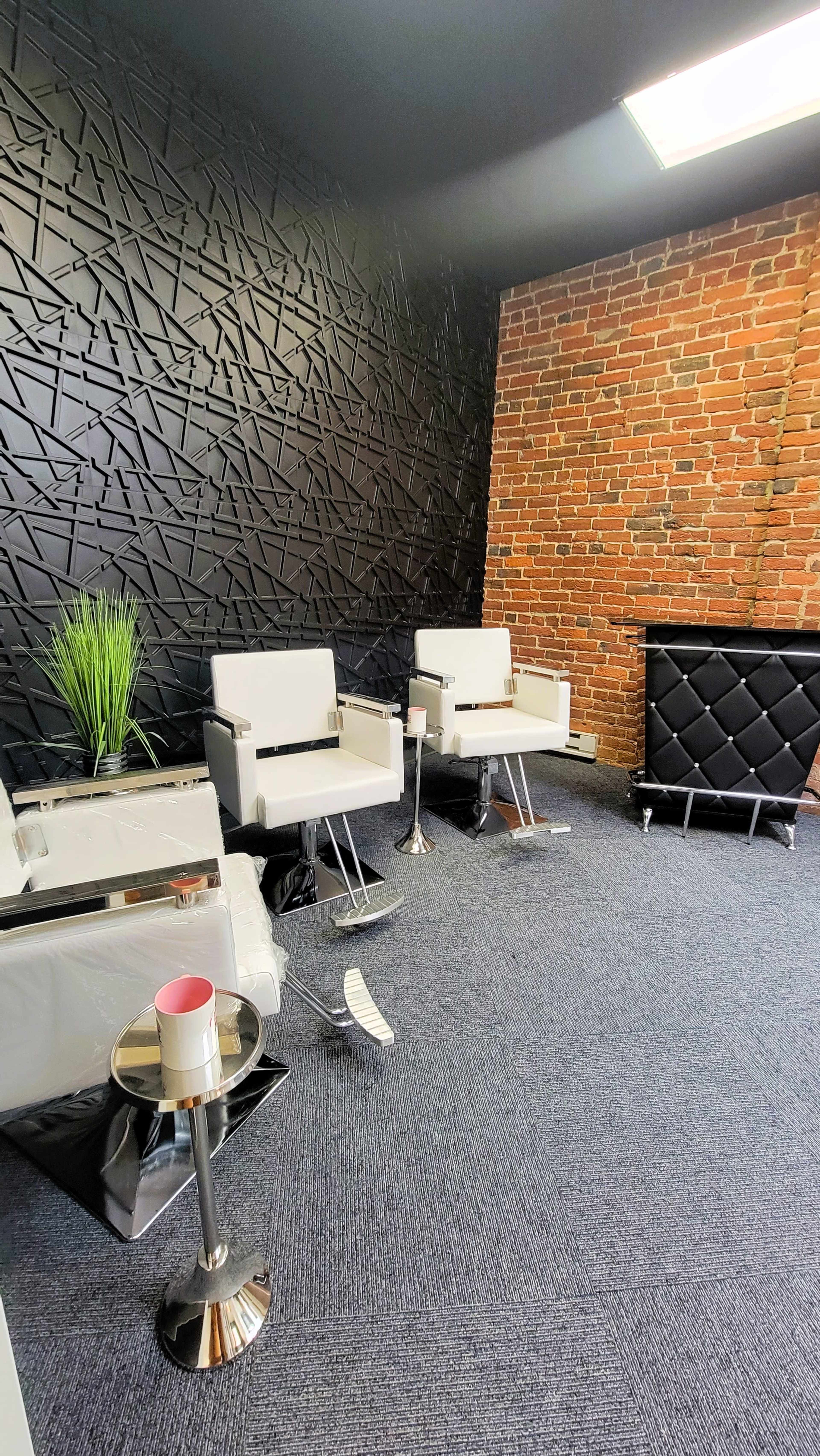A modern waiting area with two white chairs, a small glass table, a potted plant, and an accent wall made of textured black panels.