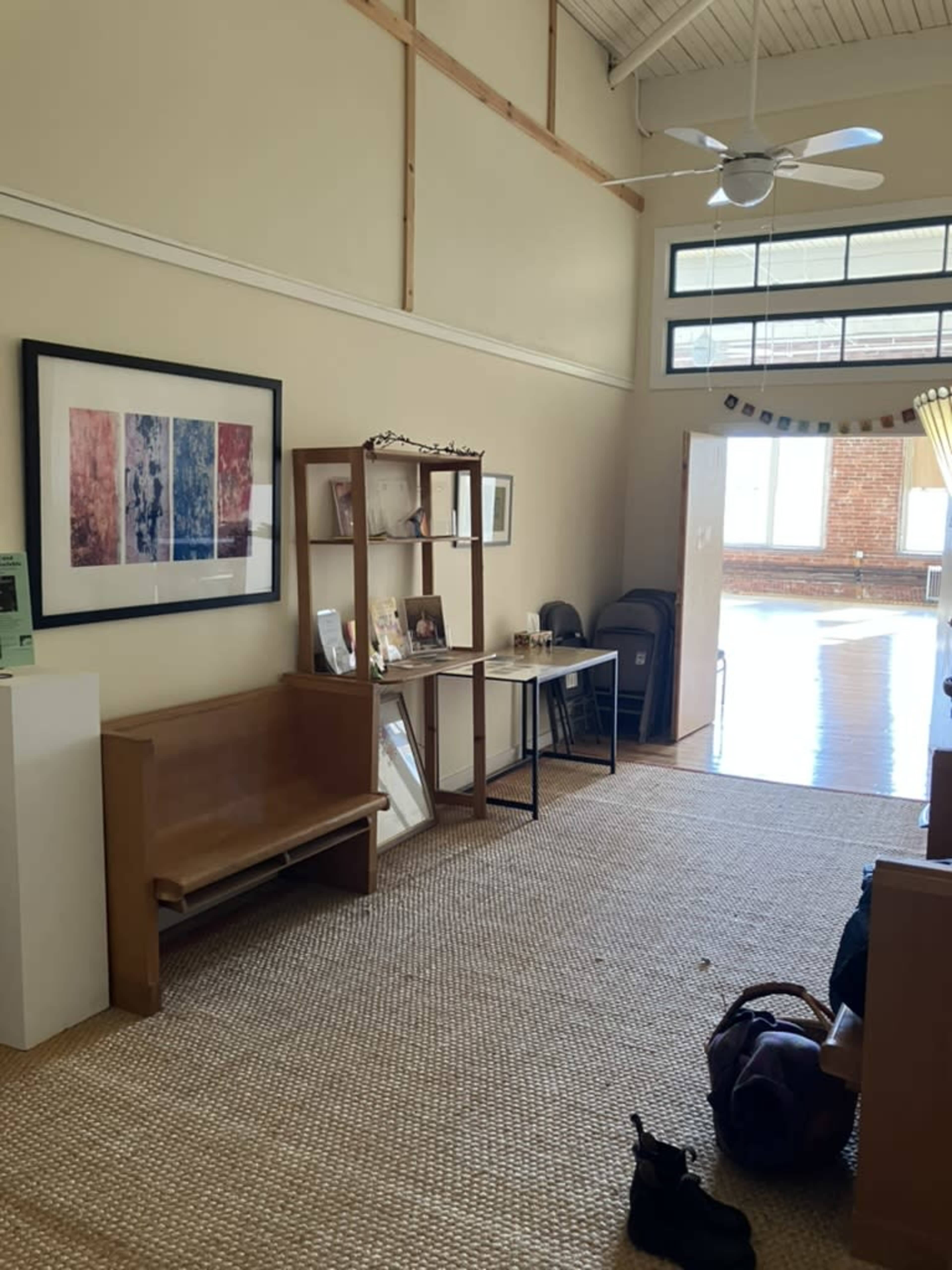 The image depicts a spacious room with a wooden bench, a display shelf containing photos, and a table, all set against a backdrop of large windows and wooden floors.
