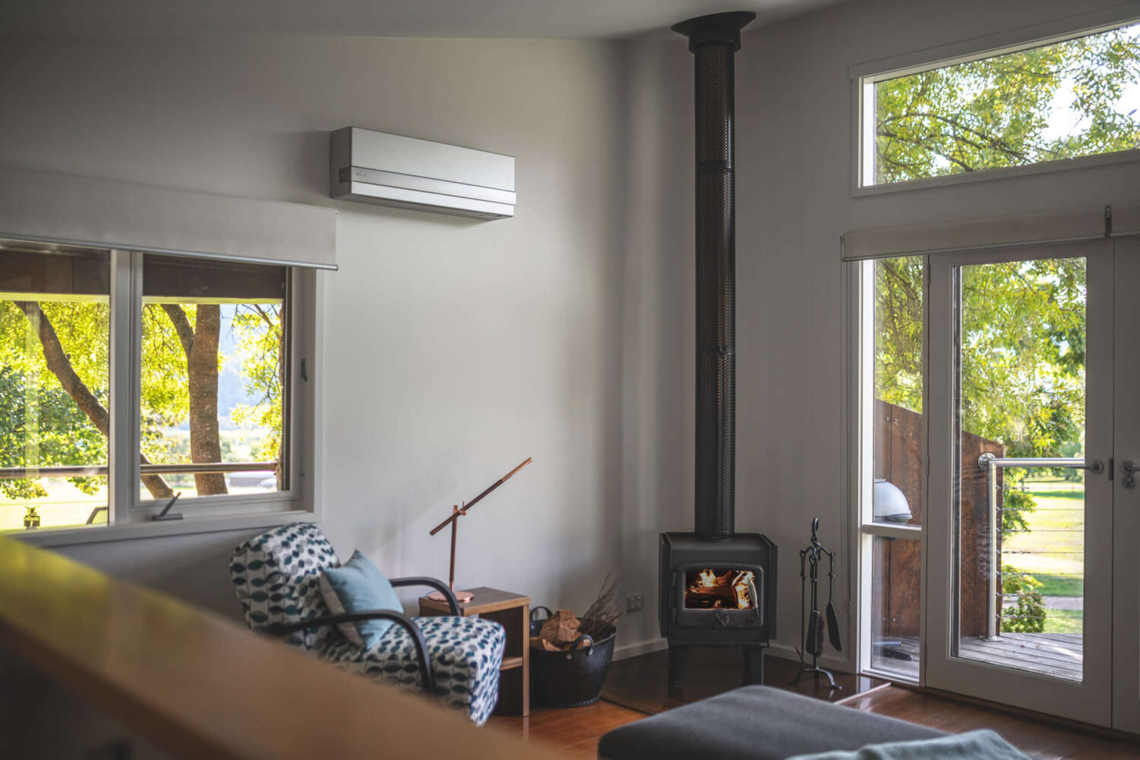 A modern living room with a fireplace, large windows allowing natural light, and a view of greenery outside.