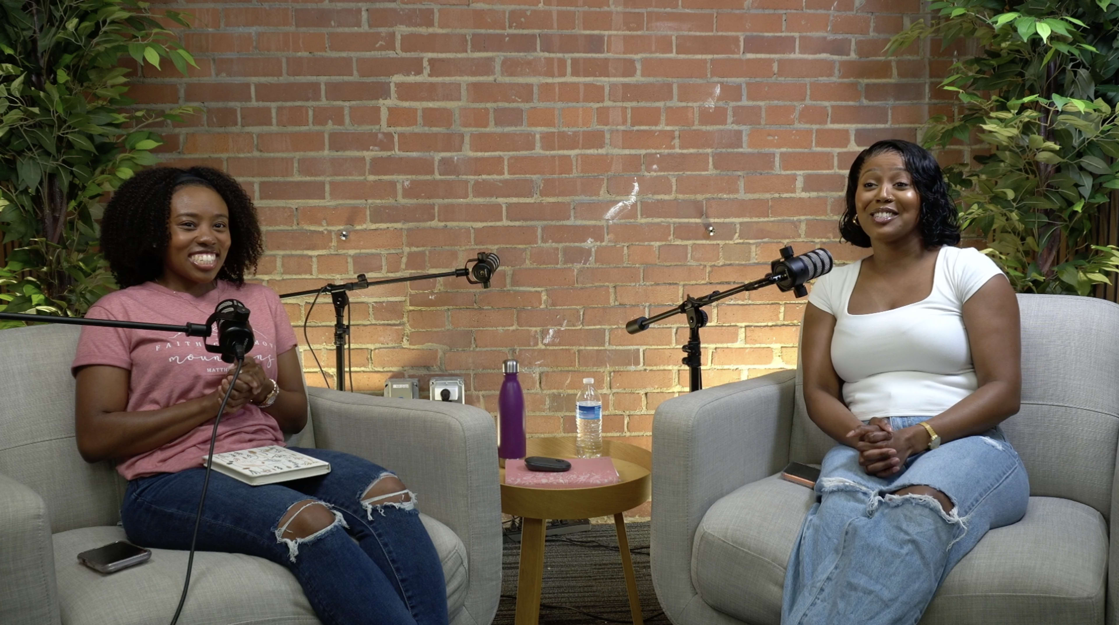 Two women sit in comfortable chairs with microphones in front of them, surrounded by greenery and a brick wall, as they engage in conversation.