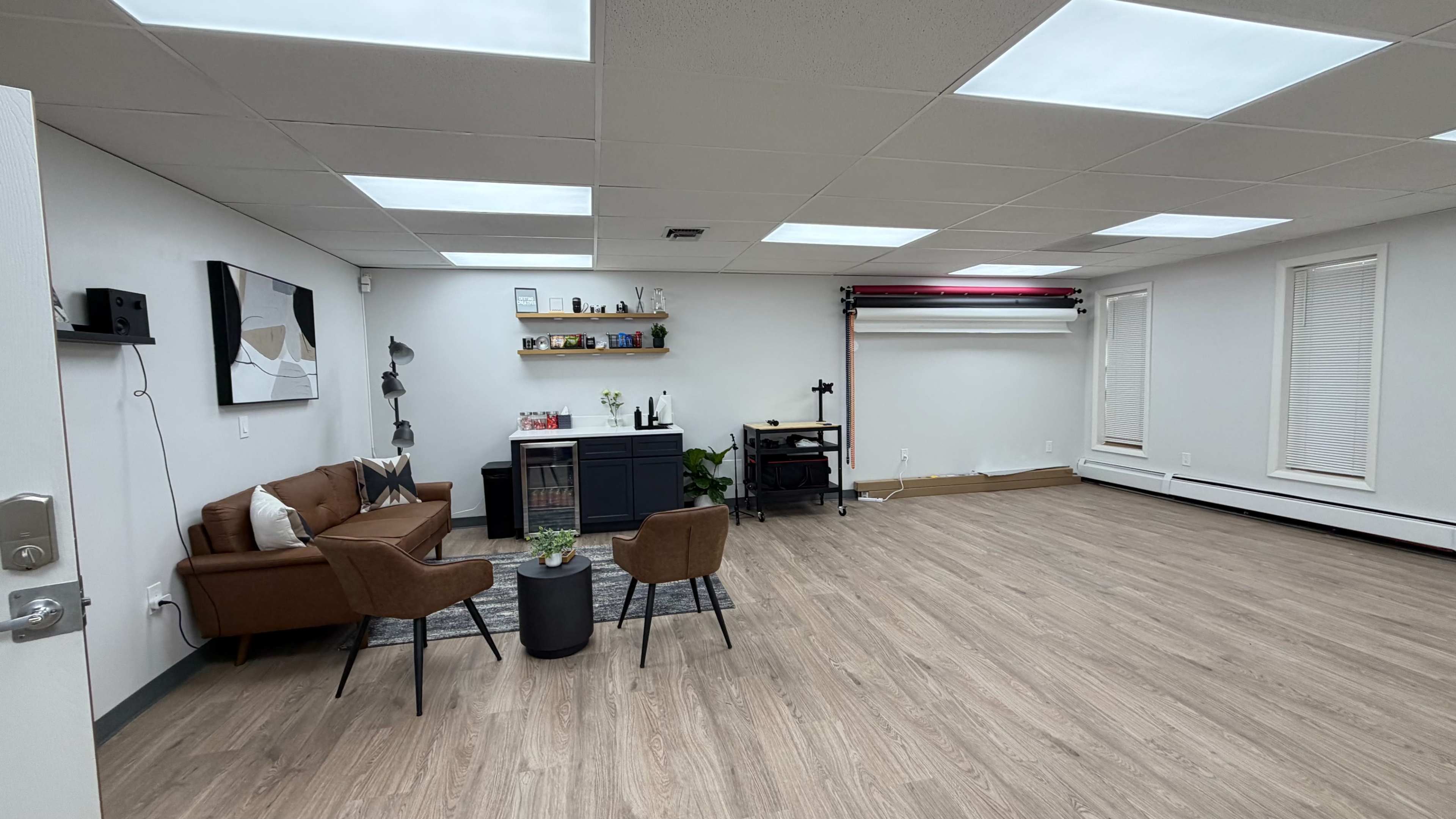 The image shows a spacious, well-lit room with a brown couch, two chairs, a small table, and minimal decor, including shelves and a cart.