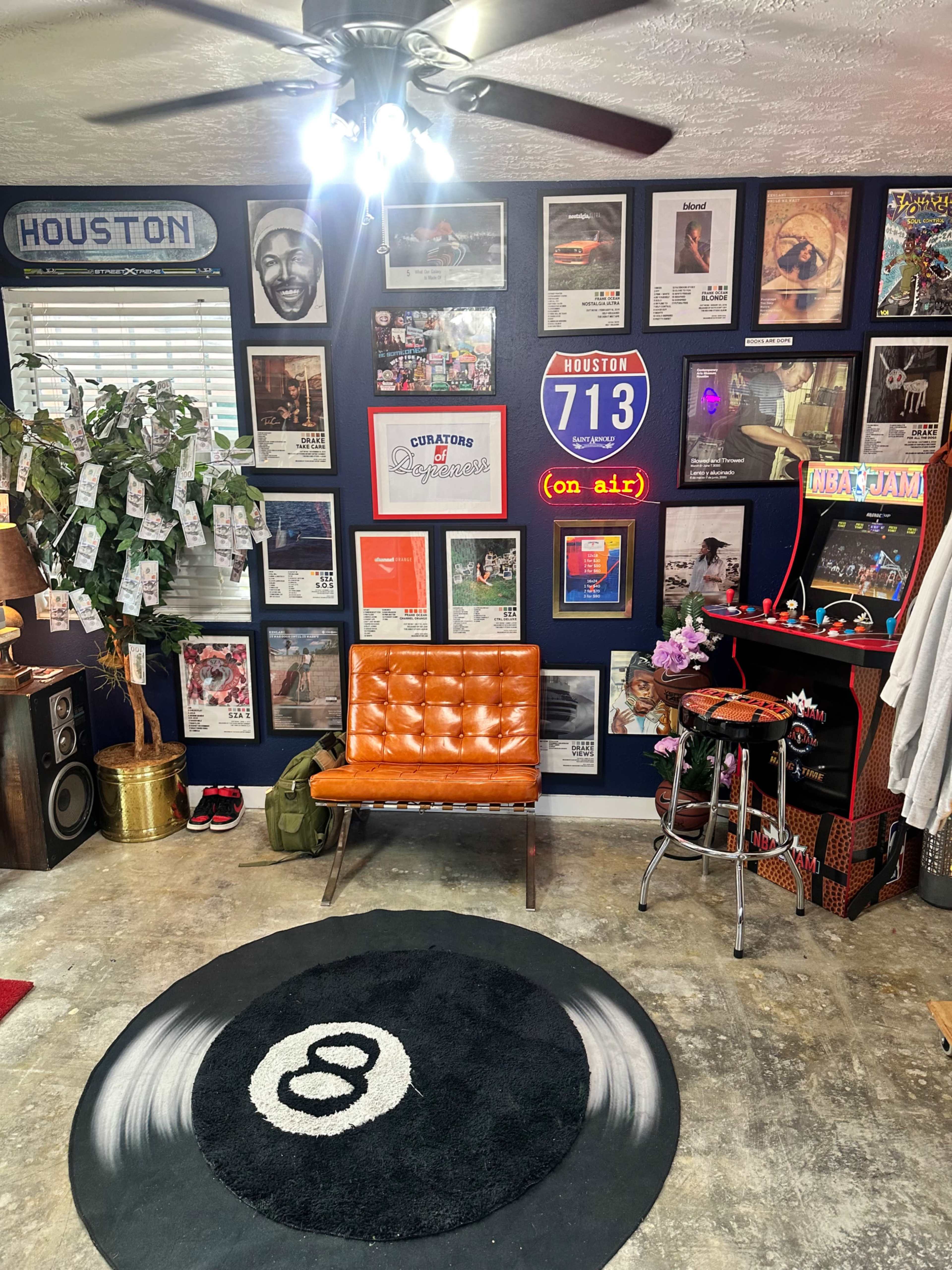 The image shows a room with a dark blue accent wall covered in framed posters and artwork, featuring an orange chair and a circular rug with an eight-ball design on the floor.