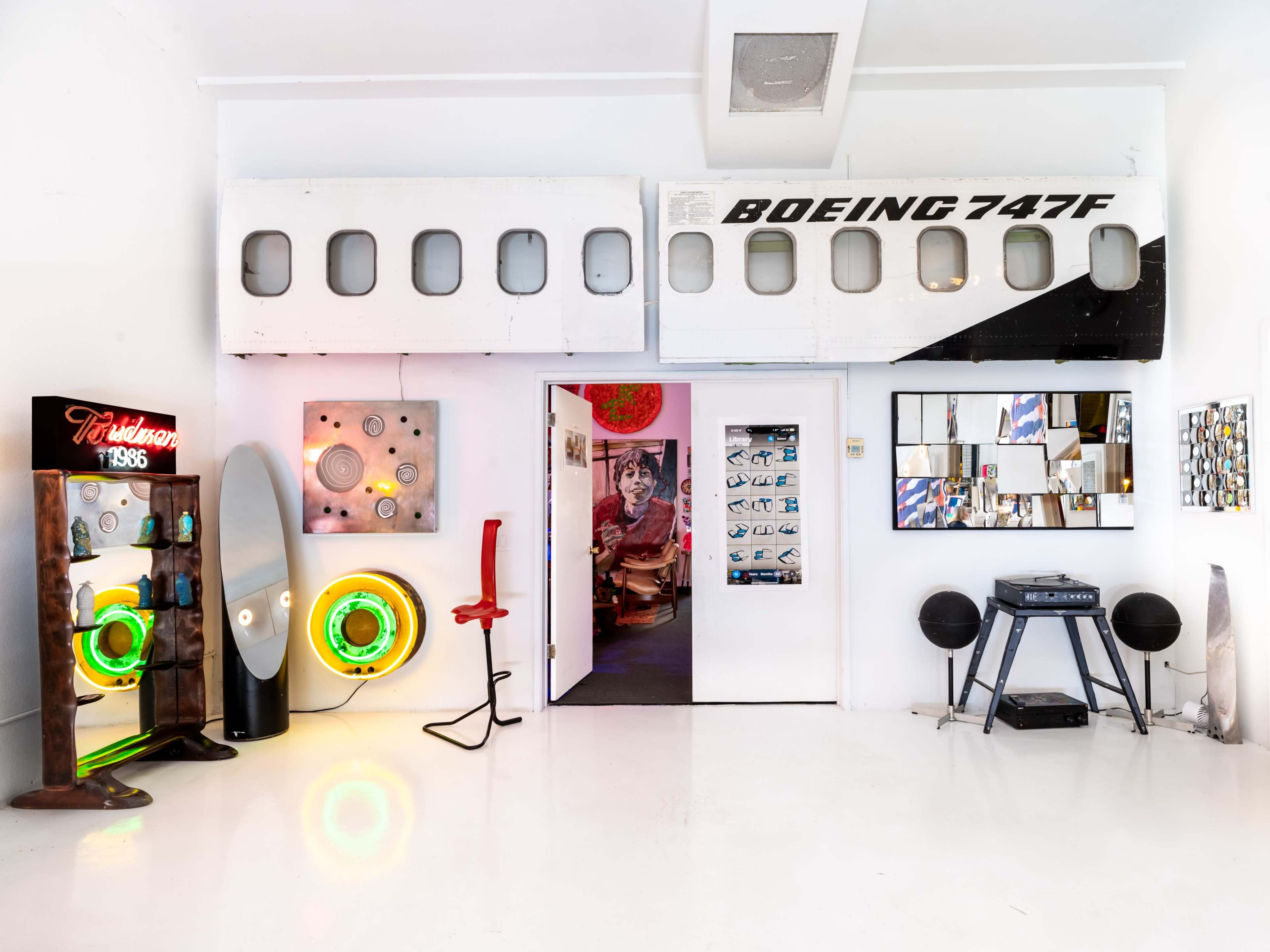 The image shows a modern art space featuring a Boeing 747 fuselage section on the wall, colorful illuminated installations, and various artworks framed on the walls.