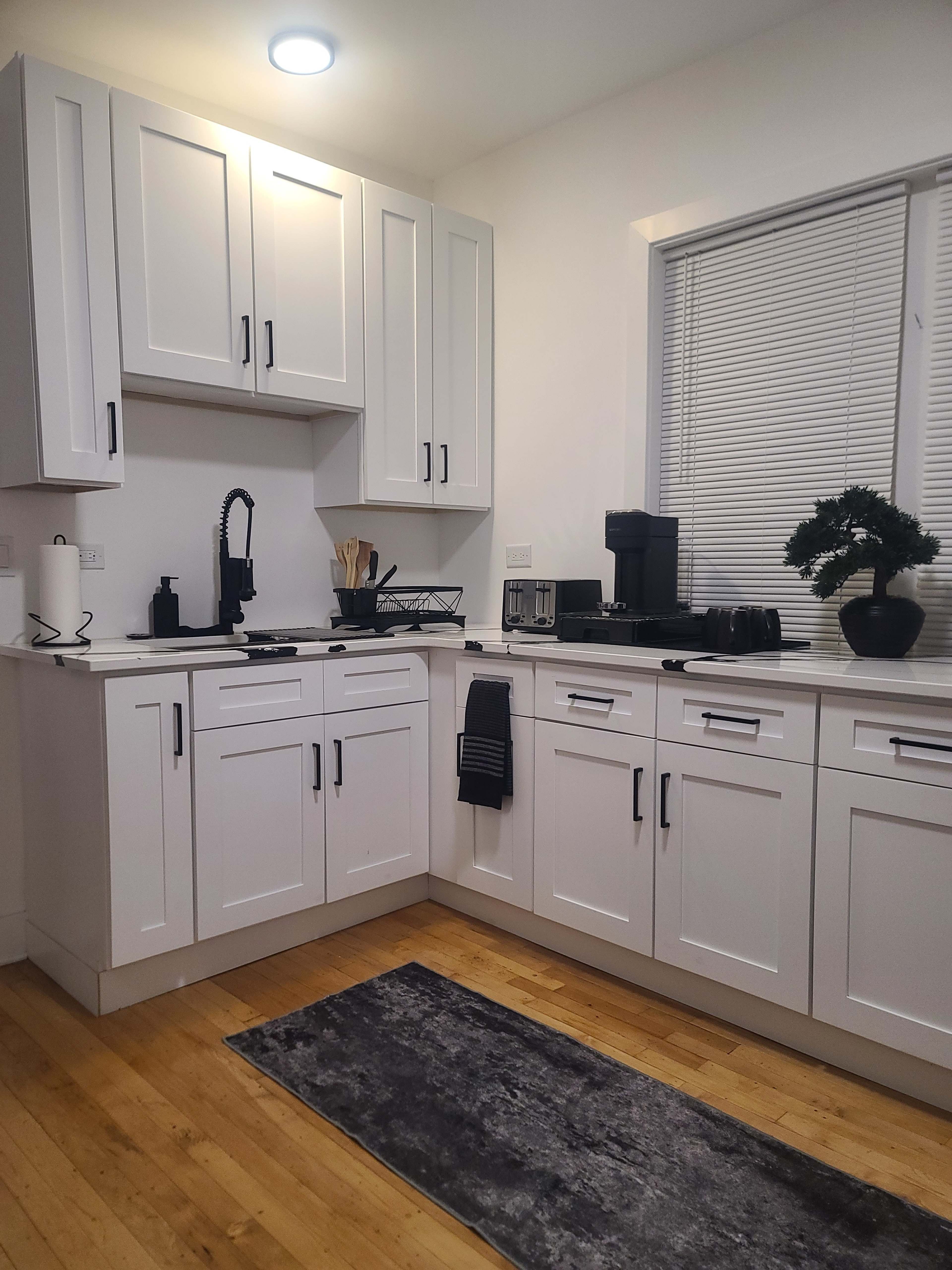 The image shows a modern kitchen featuring white cabinetry, a countertop with various kitchen appliances, and a small plant on the counter.
