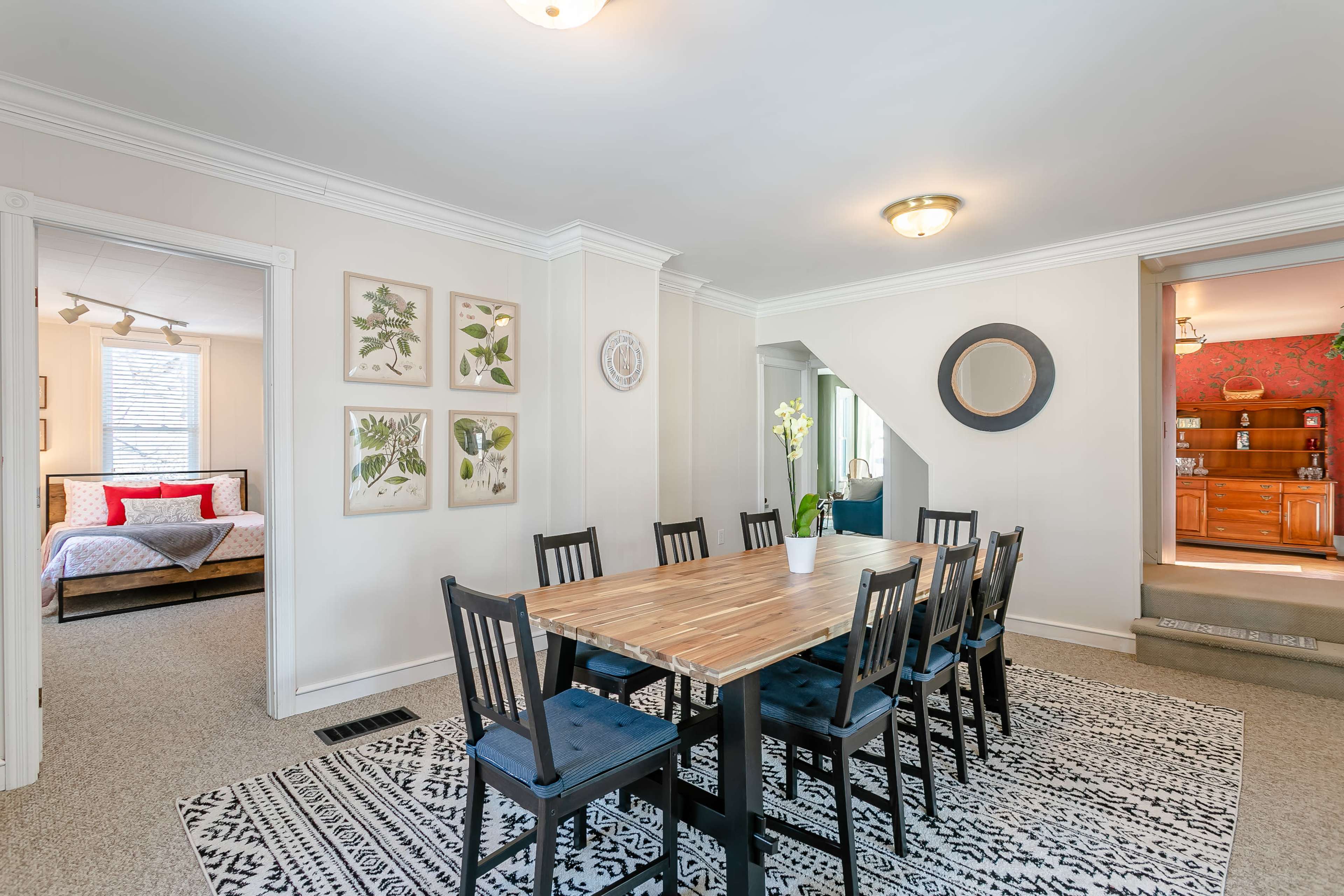 A dining room with a large wooden table and eight black chairs is bordered by two rooms, one featuring a bed and the other revealing a wooden bar area.
