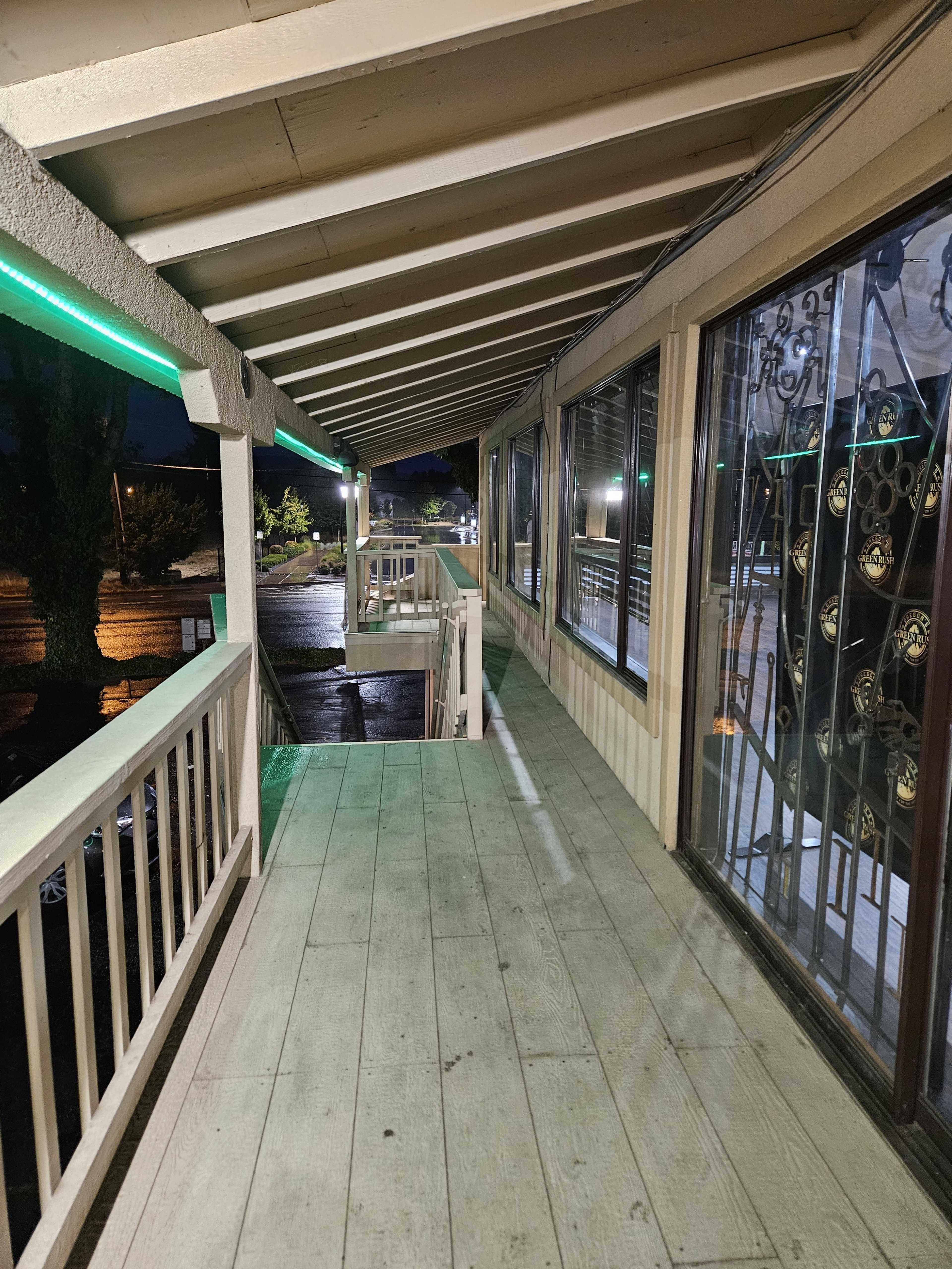 The image shows a dimly lit, covered hallway with wooden flooring and a railing, leading past large windows on one side.