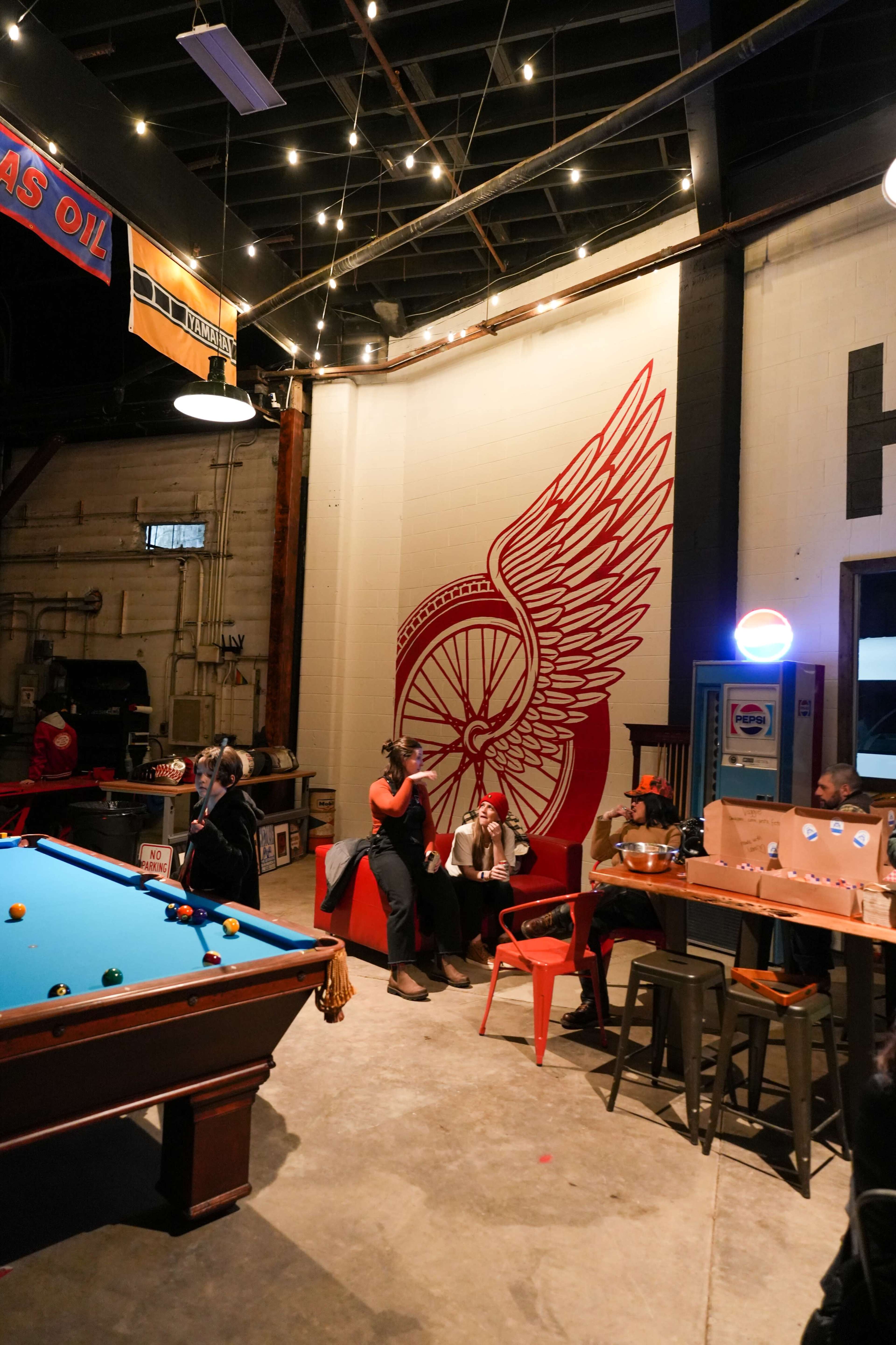 The image shows a vibrant indoor space featuring a pool table, a bar area with seating, and a large winged logo painted on the wall, with several people socializing.