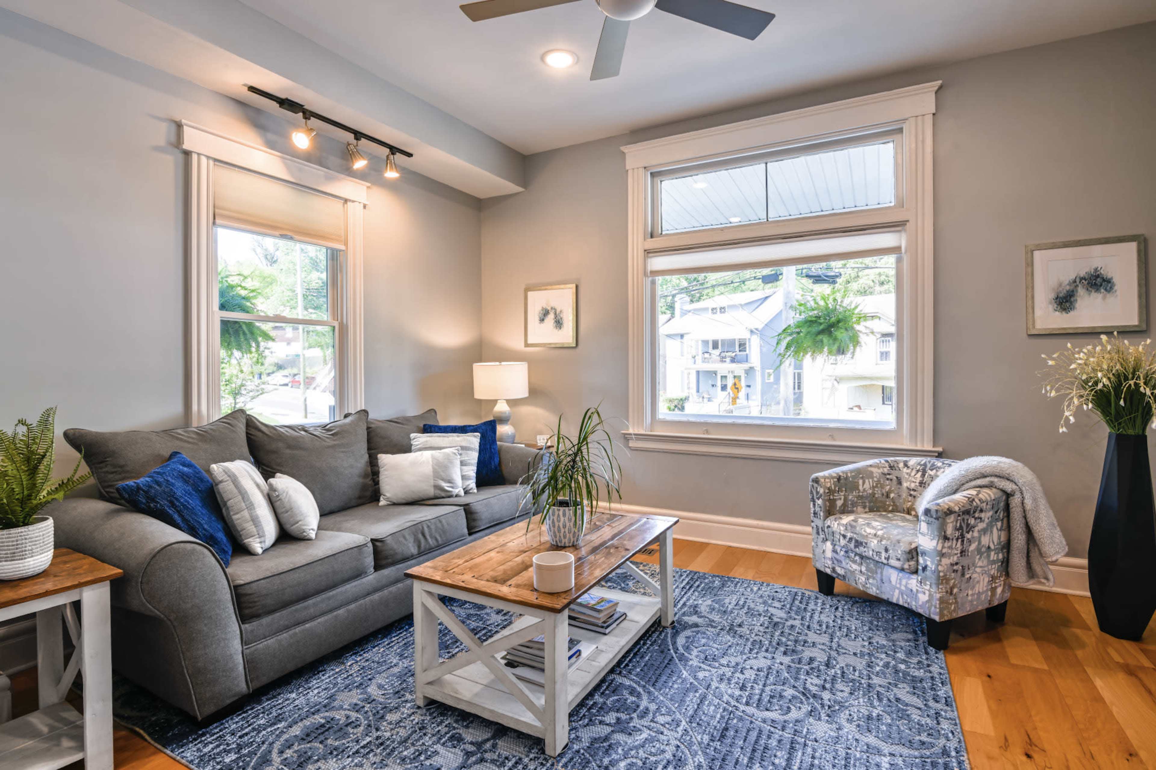 The image shows a cozy living room featuring a gray sofa, a wooden coffee table, and a patterned armchair near a large window.