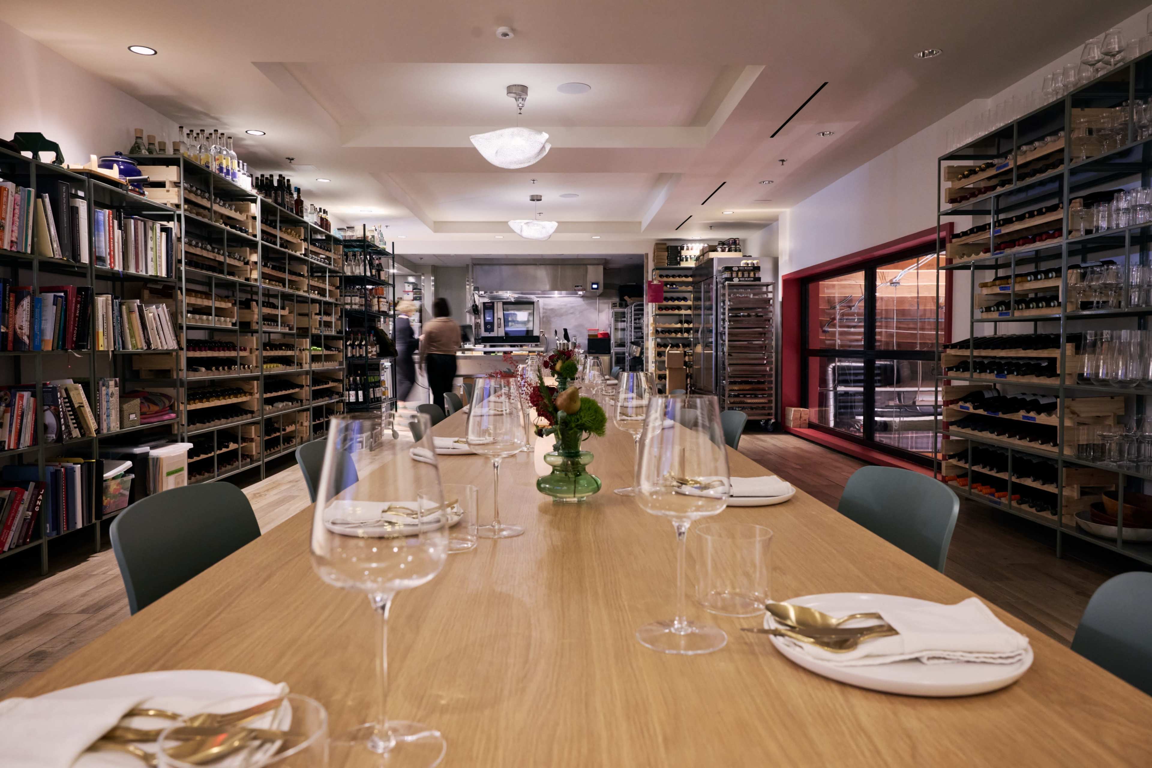 The image shows a spacious dining area with a long wooden table set for a meal, surrounded by shelves stocked with books and bottles on either side.