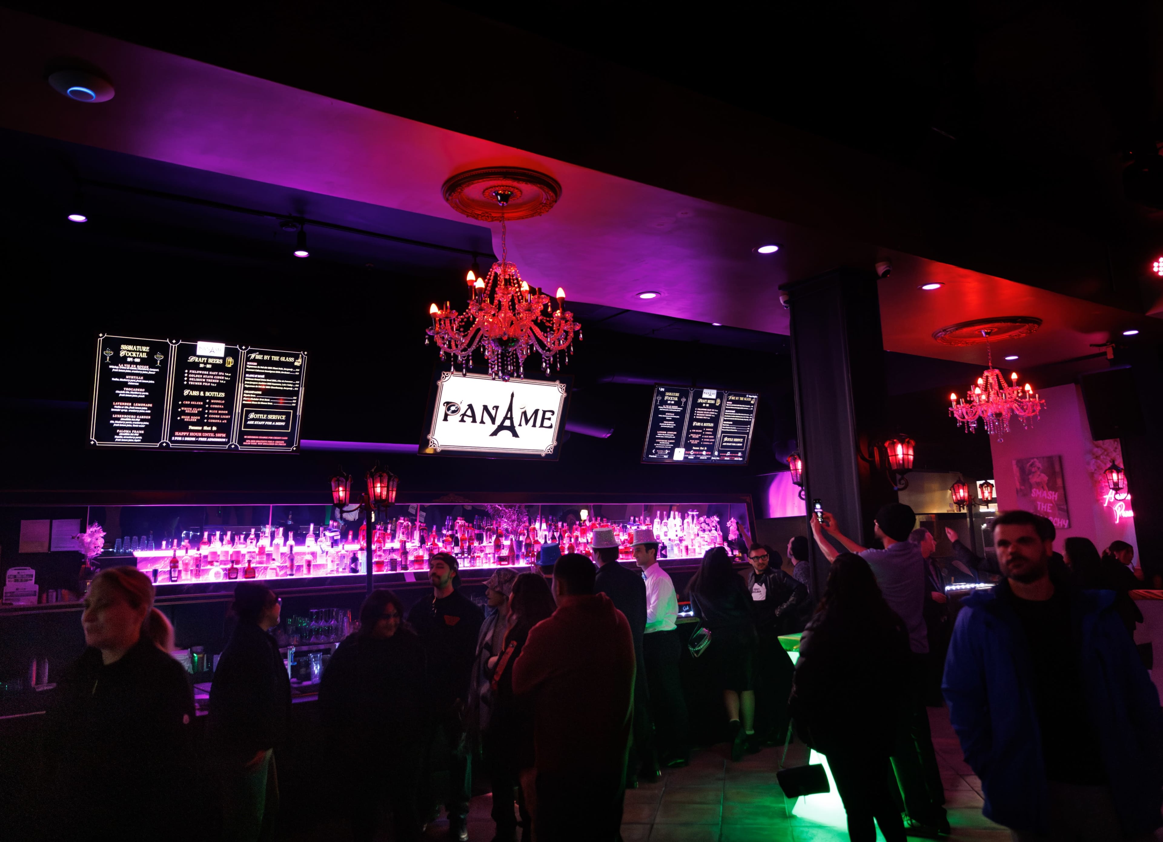 The interior of a bar features a long counter with illuminated shelves of bottles, patrons gathered around, and decorative chandeliers overhead.