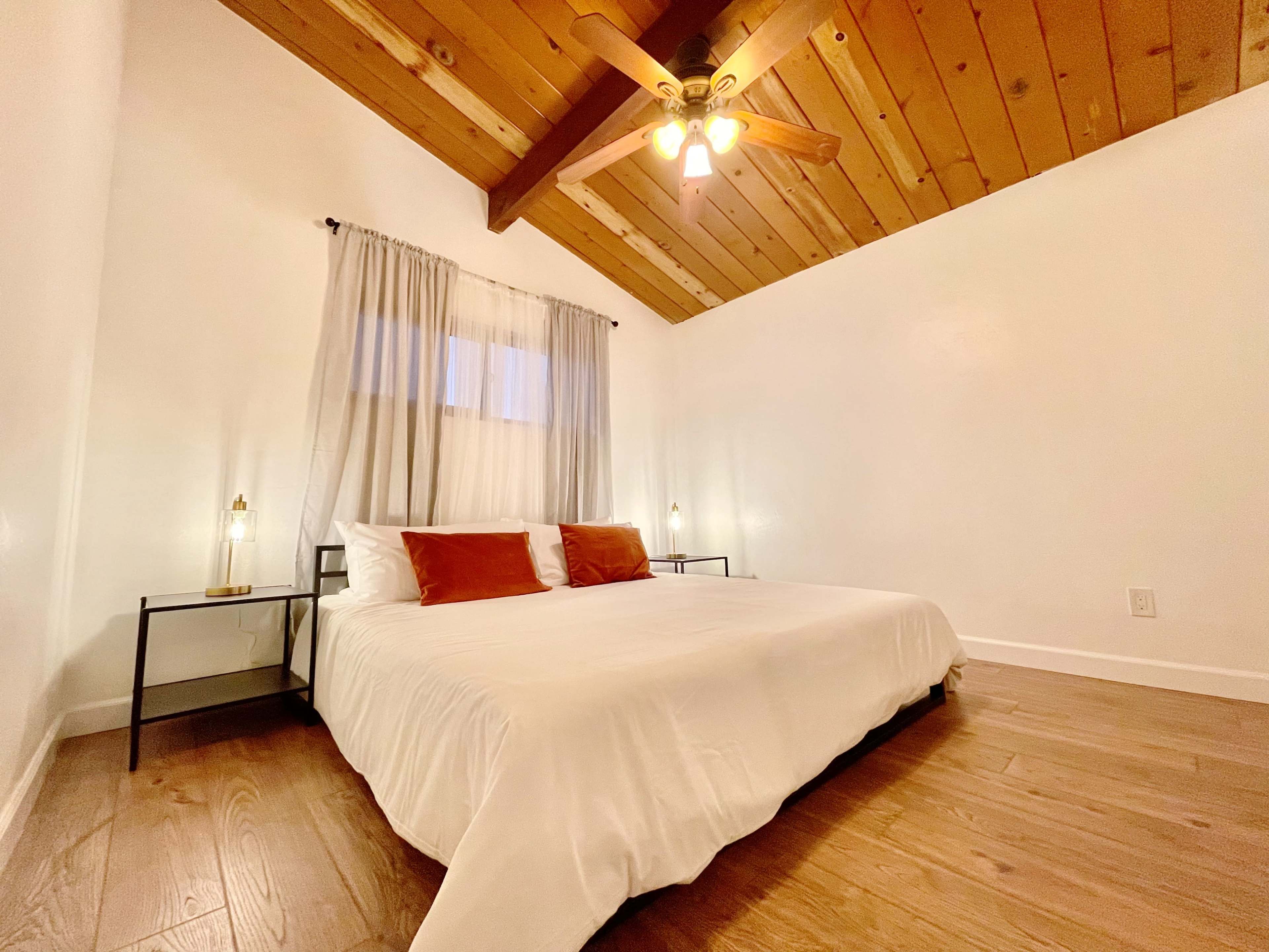 A spacious bedroom features a large bed with white linens, wooden beams in the ceiling, and soft lighting from lamps on either side.