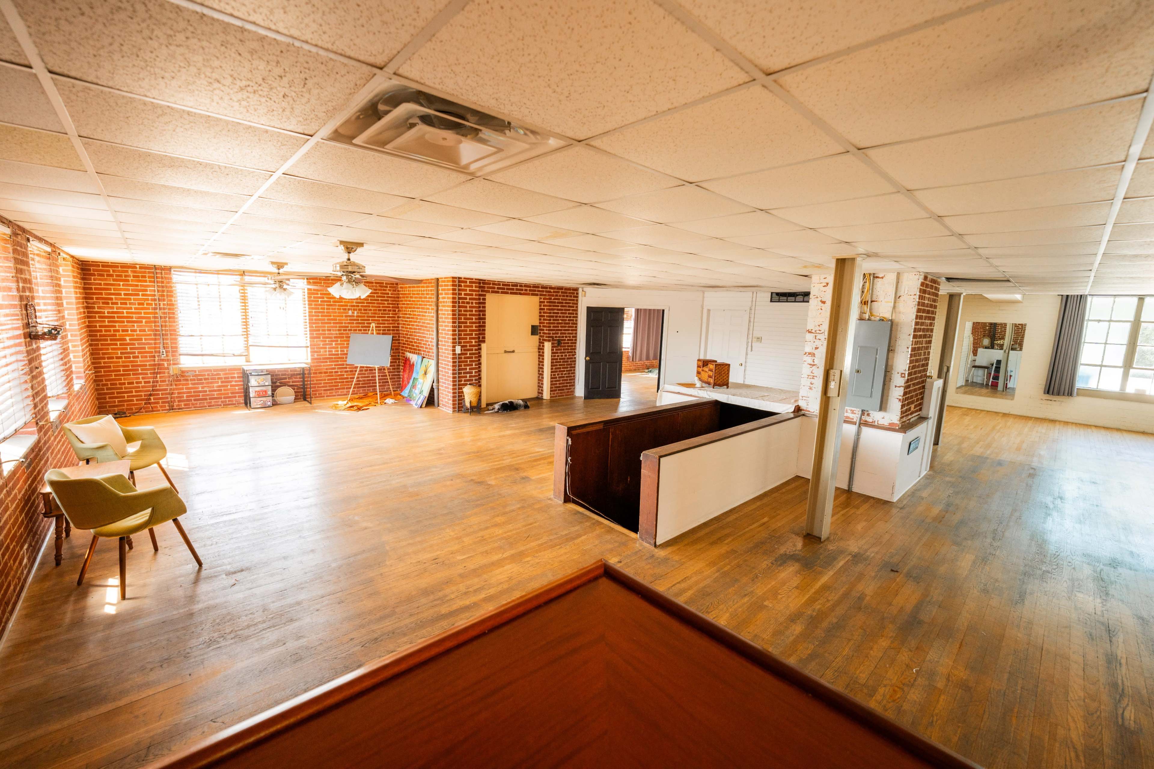 A spacious, empty room with wooden floors, brick walls, and large windows providing natural light.