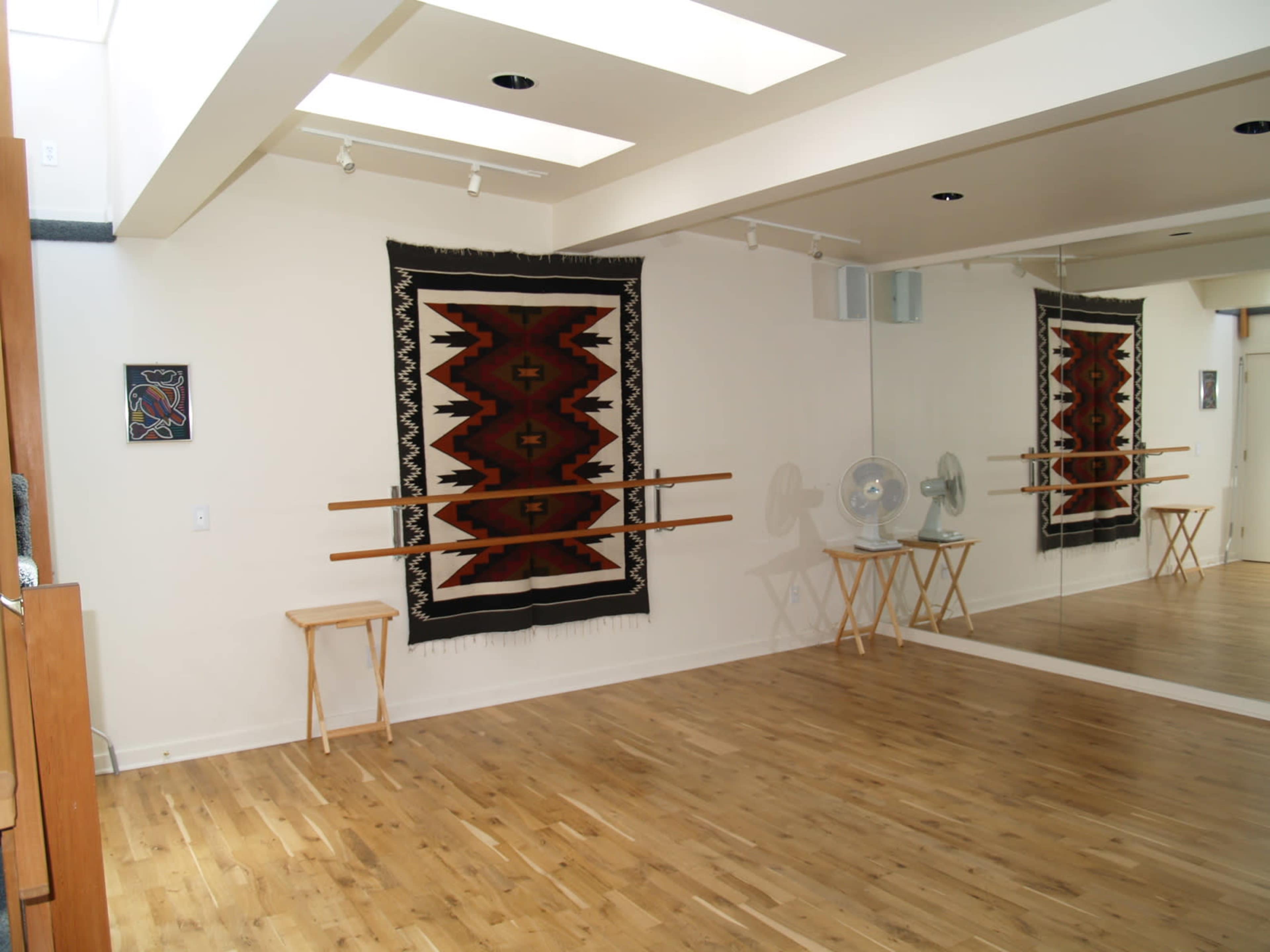 A dance studio with a wooden floor, a large mirror on one wall, and an artistic tapestry hanging on the opposite wall.