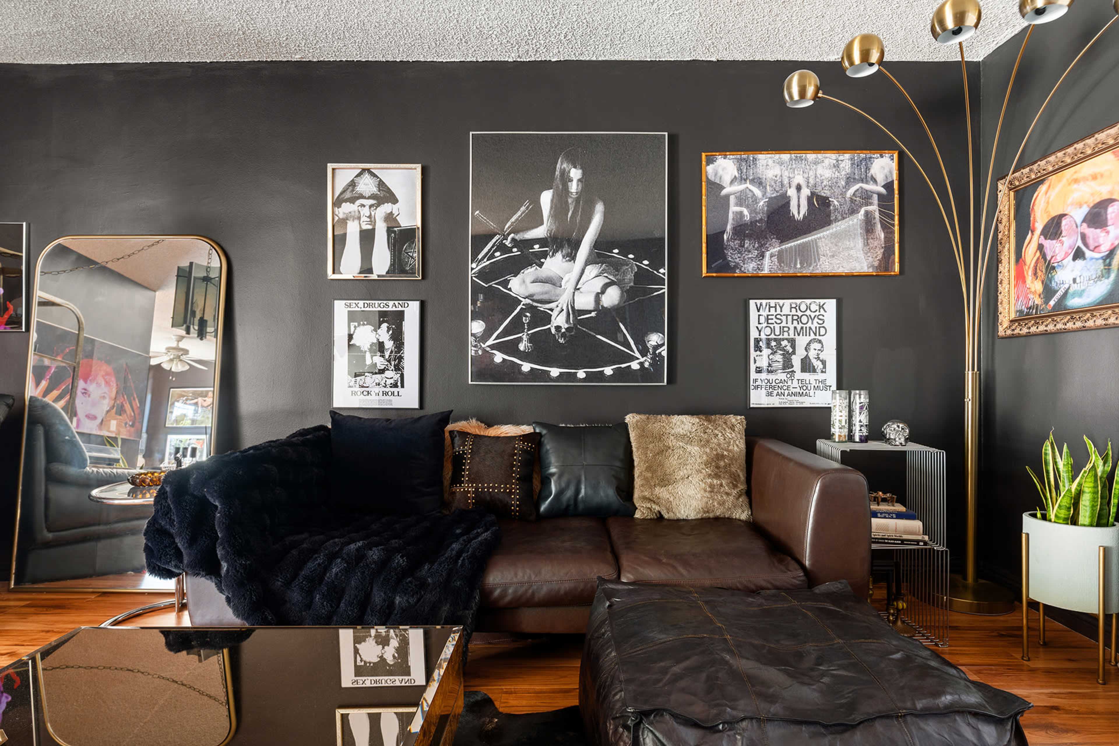 The image shows a modern living room with dark walls, featuring a mix of framed artwork, a large mirror, and a sectional sofa adorned with various textured pillows and throws.