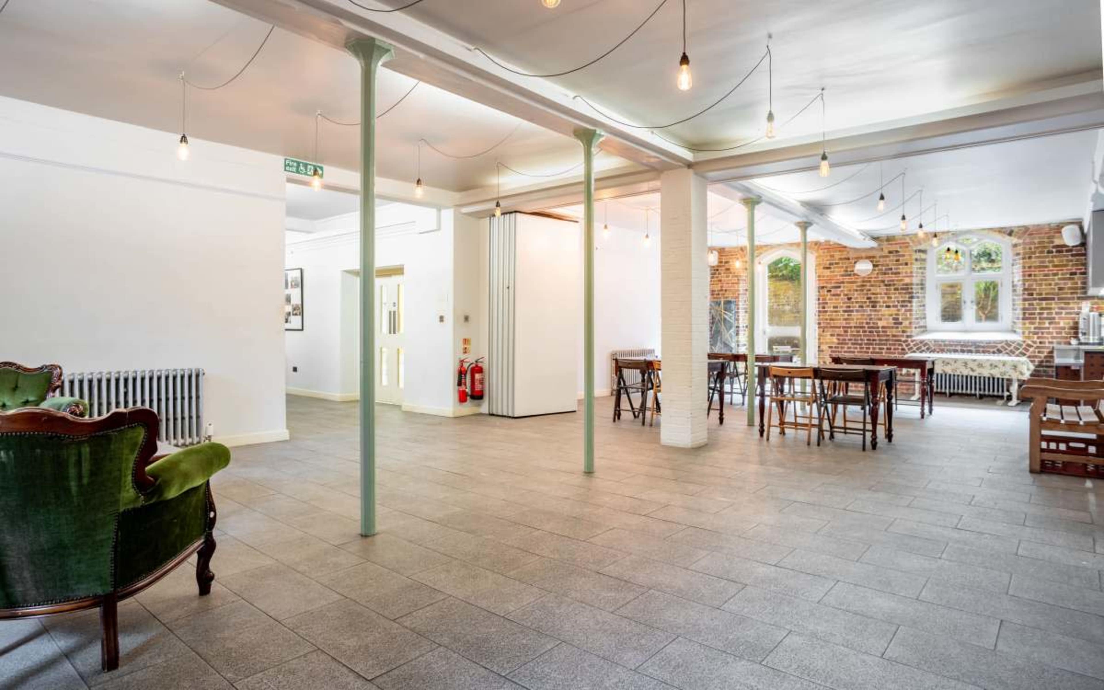 A spacious, well-lit room with a mix of seating areas, including a green upholstered armchair and several tables arranged throughout.