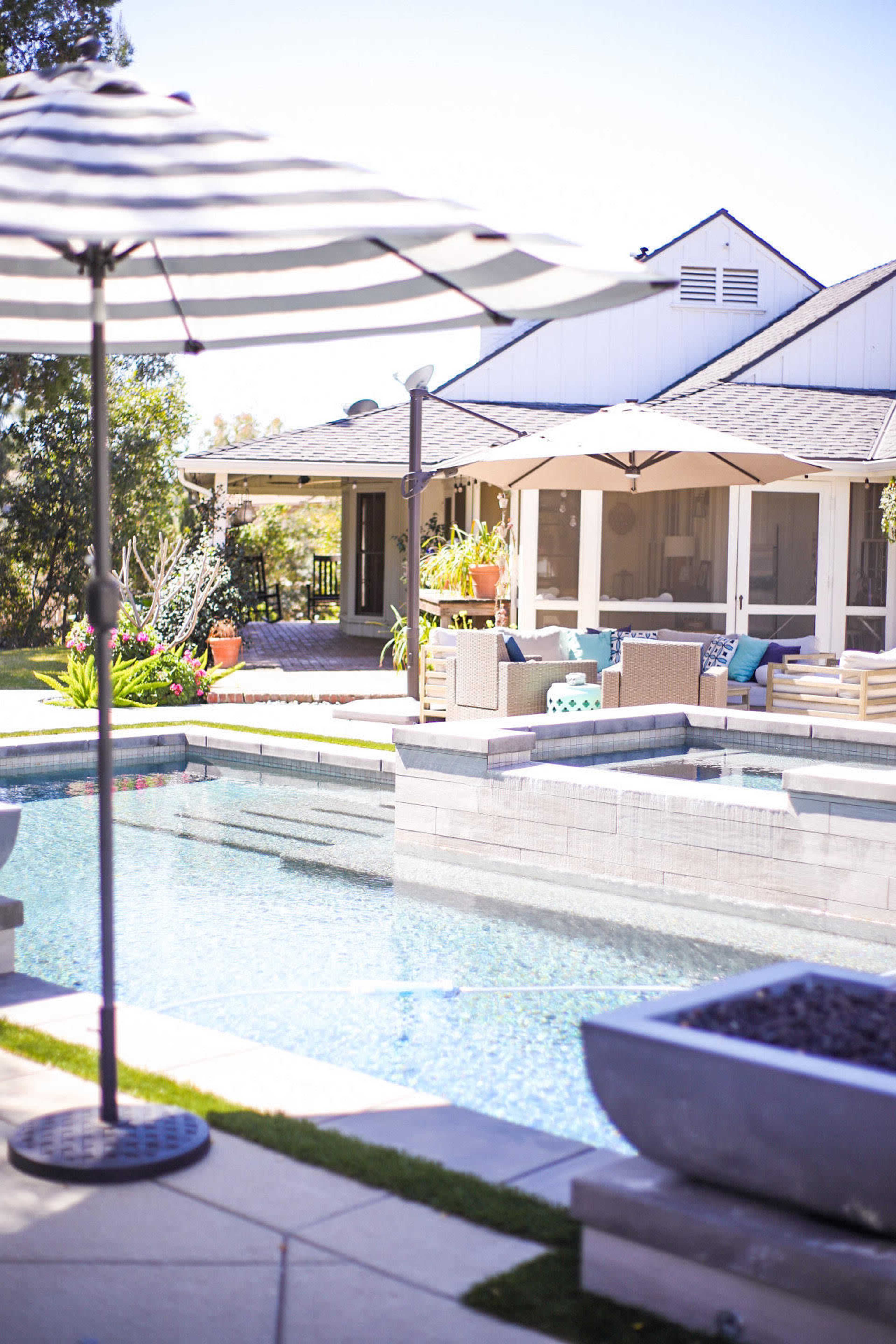 A backyard pool area with a striped umbrella, lounge furniture, and a house in the background surrounded by greenery.