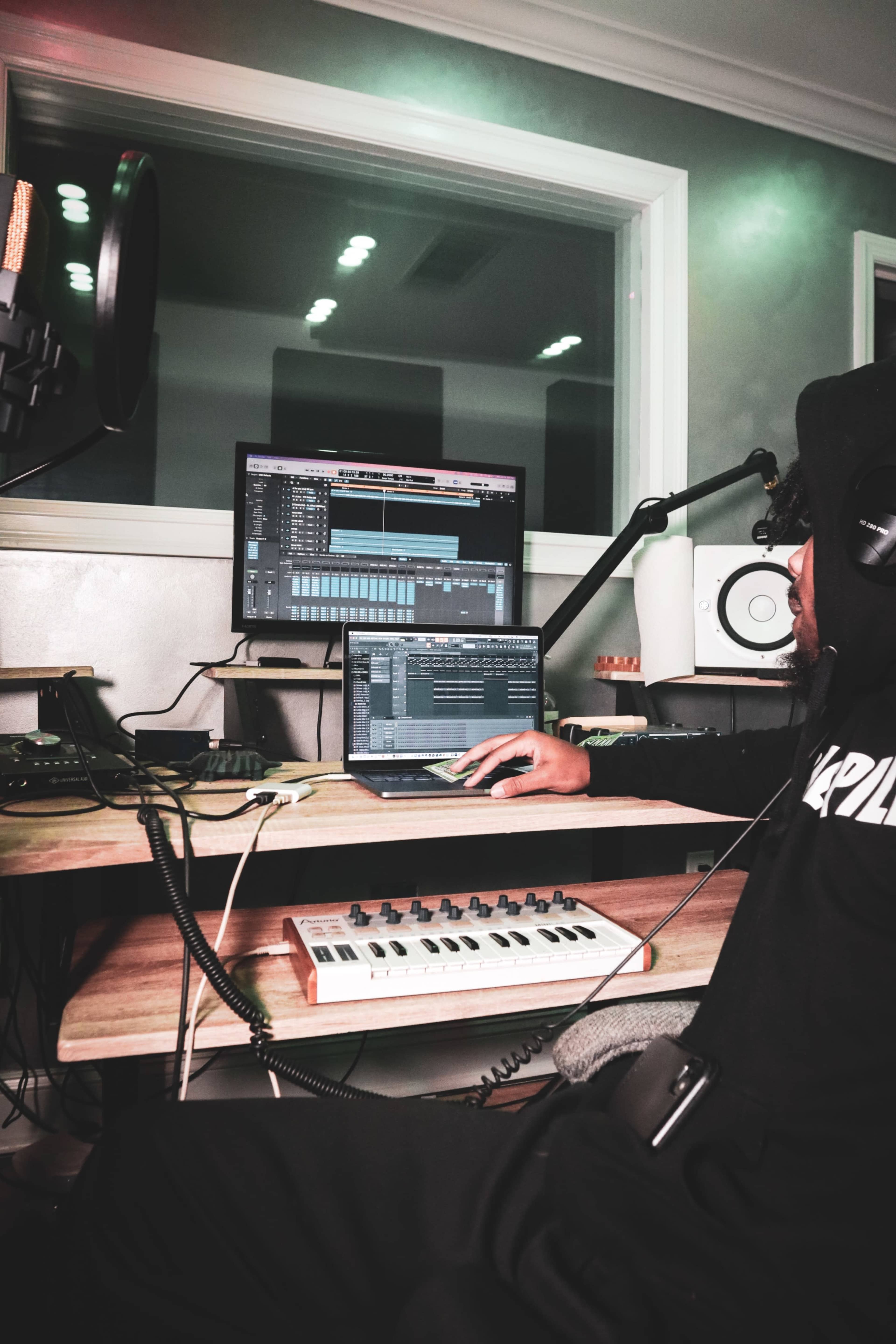A person is sitting at a desk in a music studio, using a laptop and a MIDI keyboard while working on a digital audio workstation.