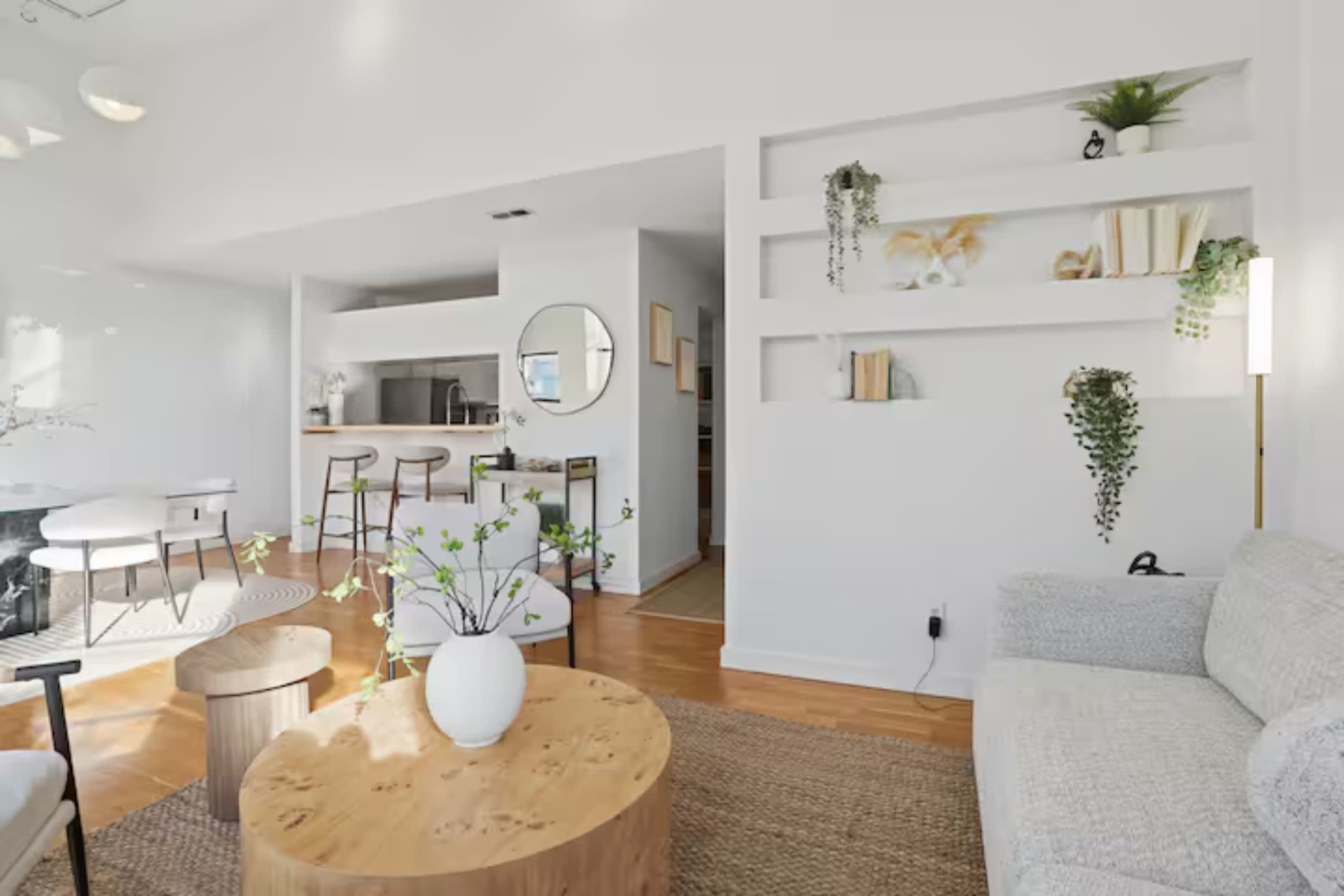 The room features a bright, open layout with a sofa, a wooden coffee table, a dining area, and shelves adorned with plants and decor.