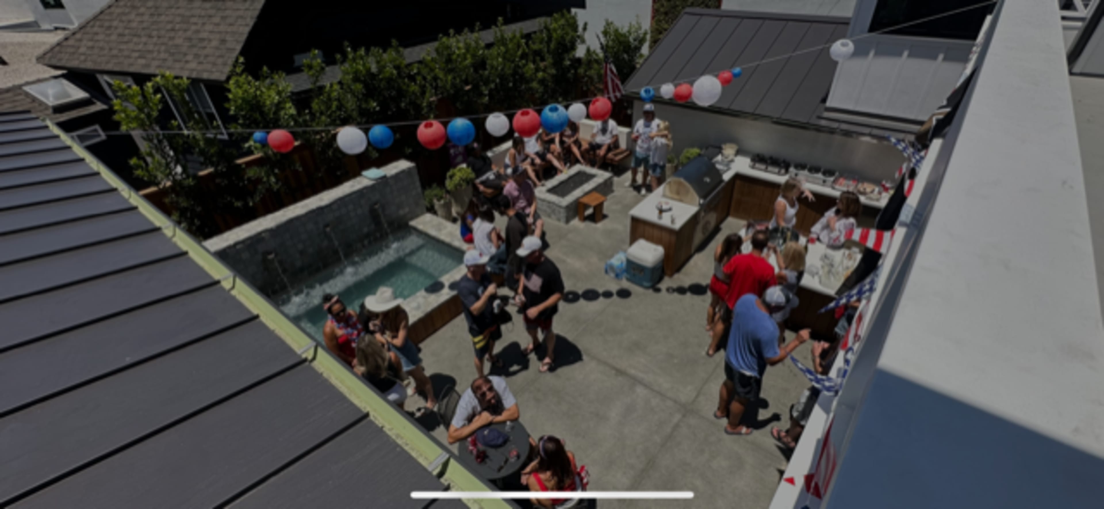 A large gathering of people is taking place in an outdoor space with a pool and decorated tables, featuring red, white, and blue balloons.