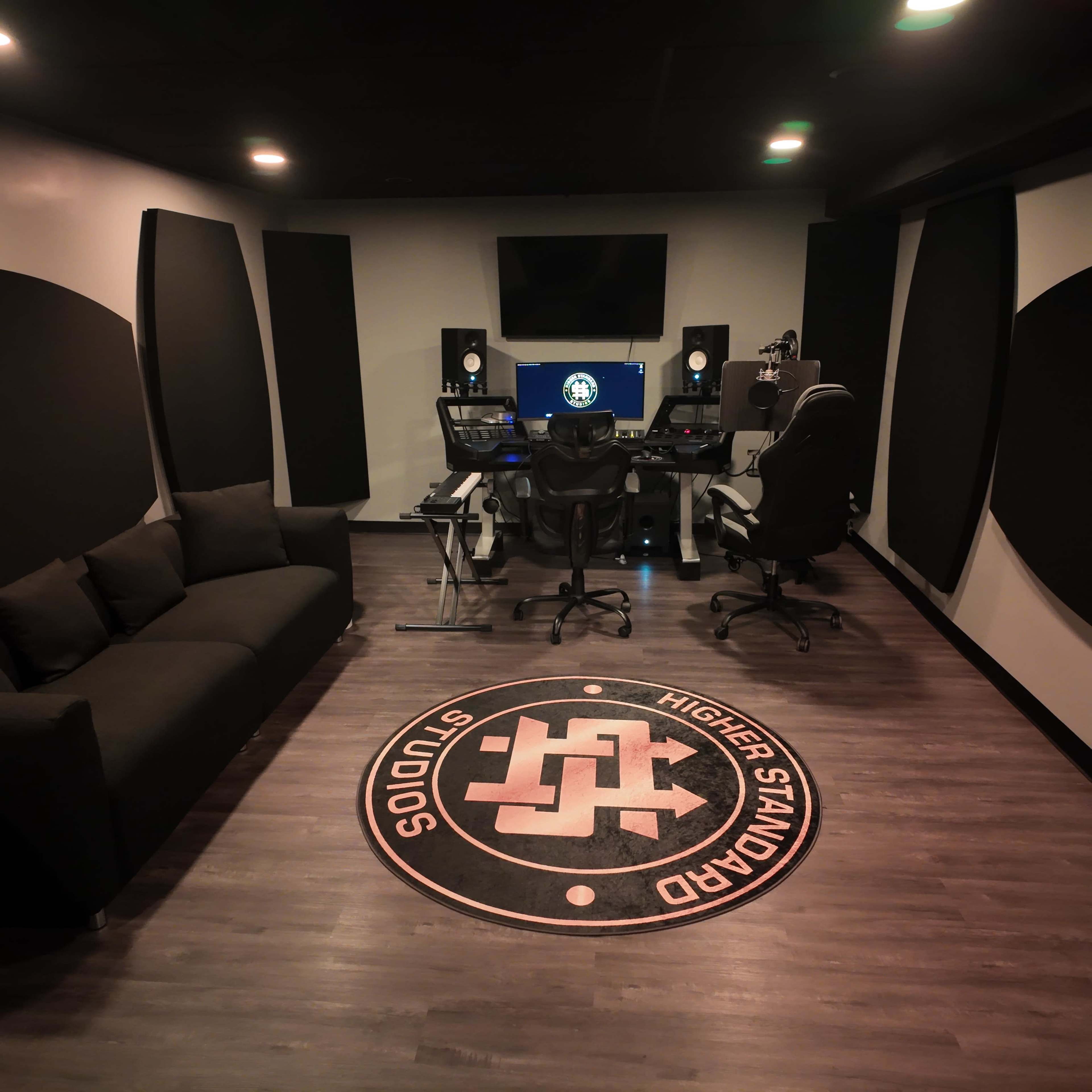 The image shows a modern music studio setup with a desk containing audio equipment, a large monitor, and a comfortable seating area with a black couch.