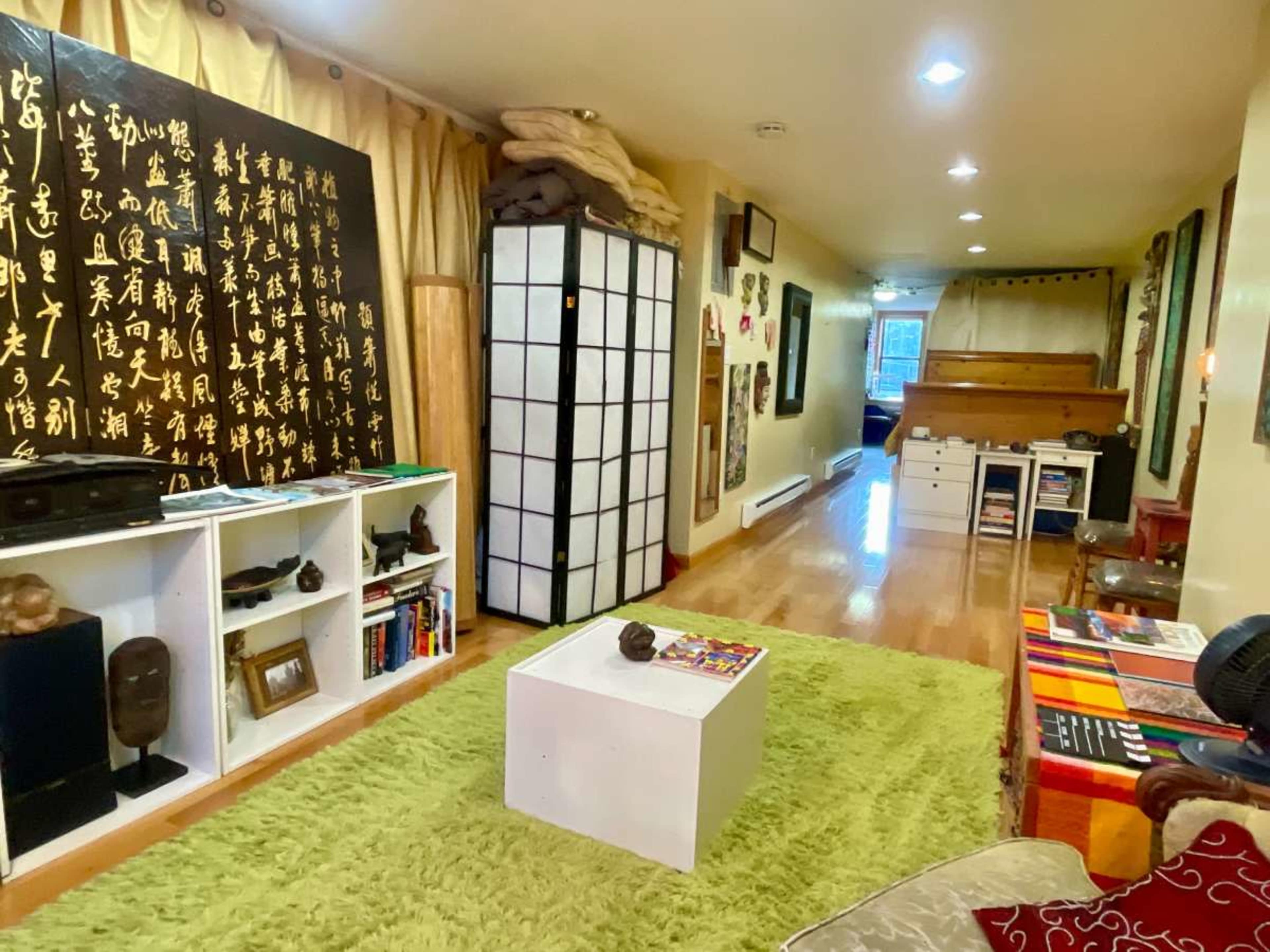 The image shows a long, narrow room featuring a green rug, a white coffee table, and walls adorned with various artworks and decorations, including a black panel with calligraphy.