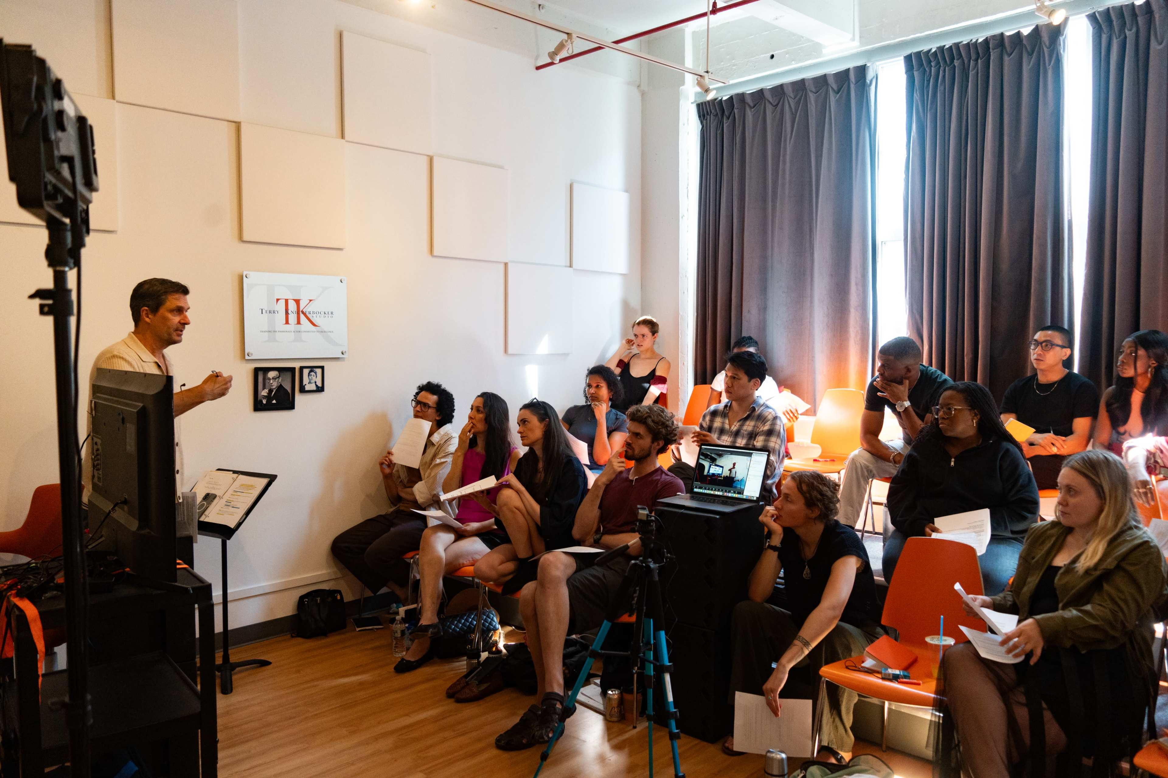 A teacher instructs a group of students seated in a classroom with large windows and modern decor.