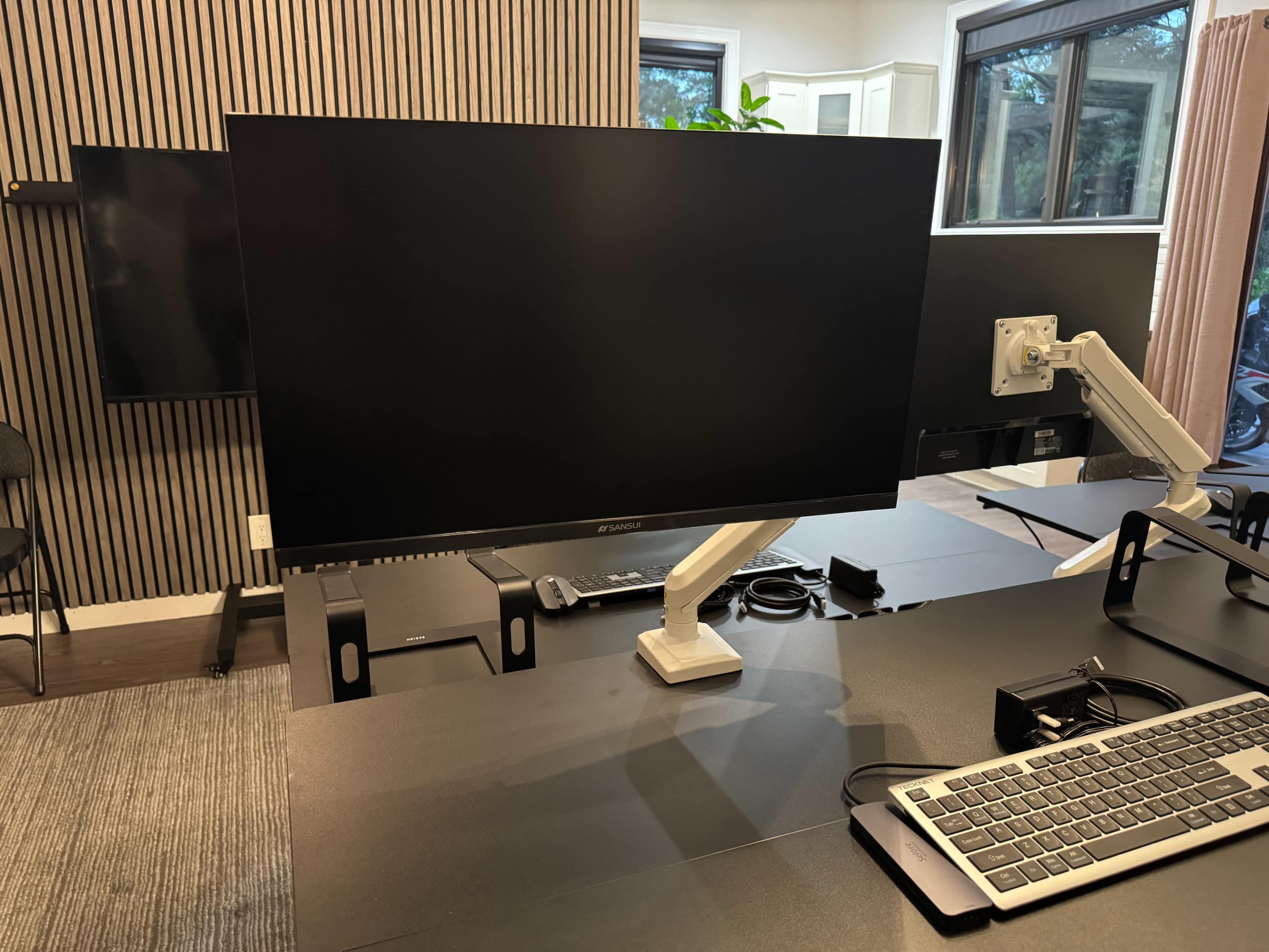 The image shows a modern workspace featuring two large monitors on adjustable arms, a keyboard, and a desk setup with a minimalist design.