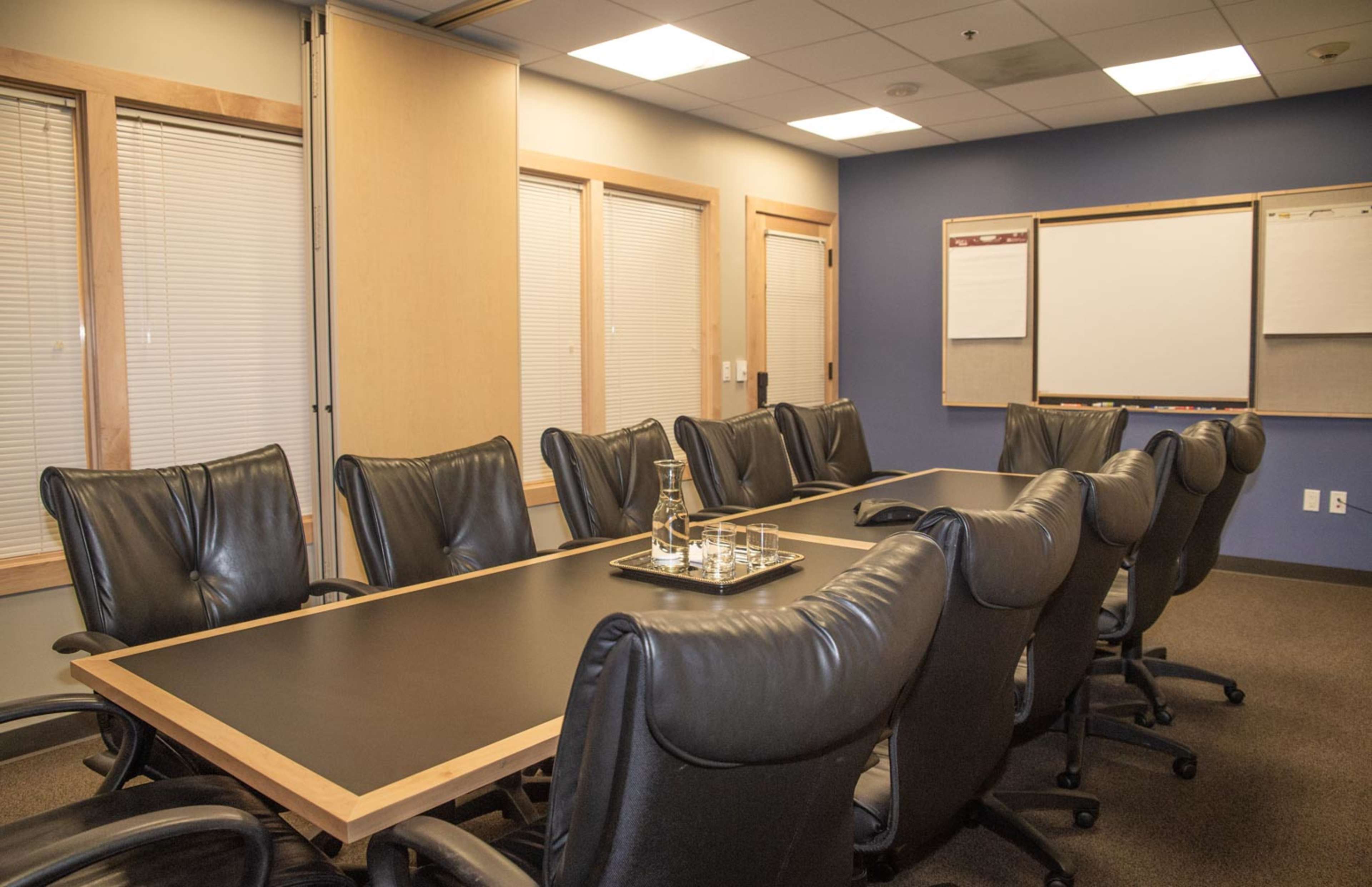 A conference room with a large wooden table surrounded by black leather chairs, featuring a whiteboard and presentation boards on the walls.