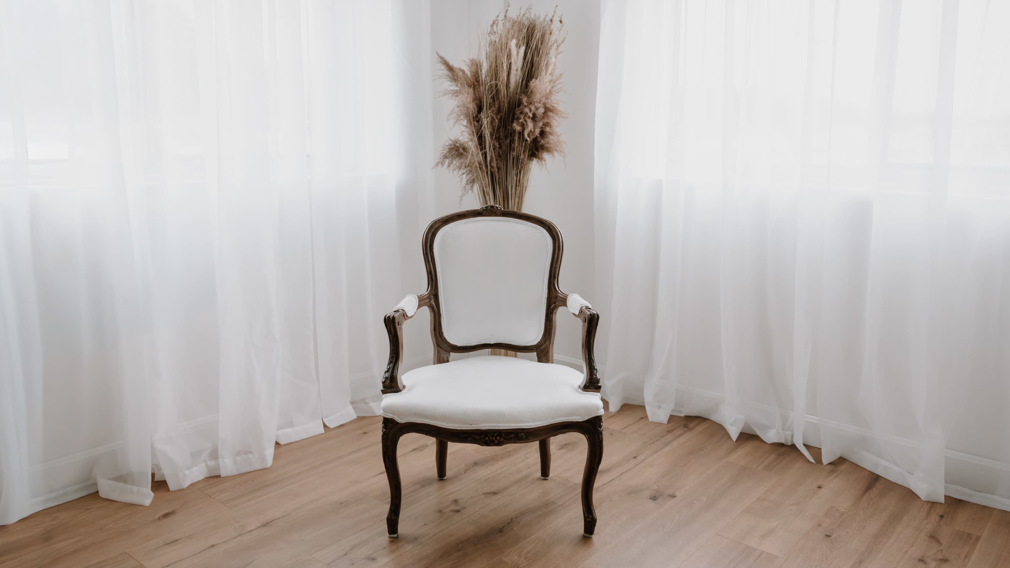 A vintage chair with a white cushion is positioned in front of sheer white curtains and a decorative arrangement of dried plants.
