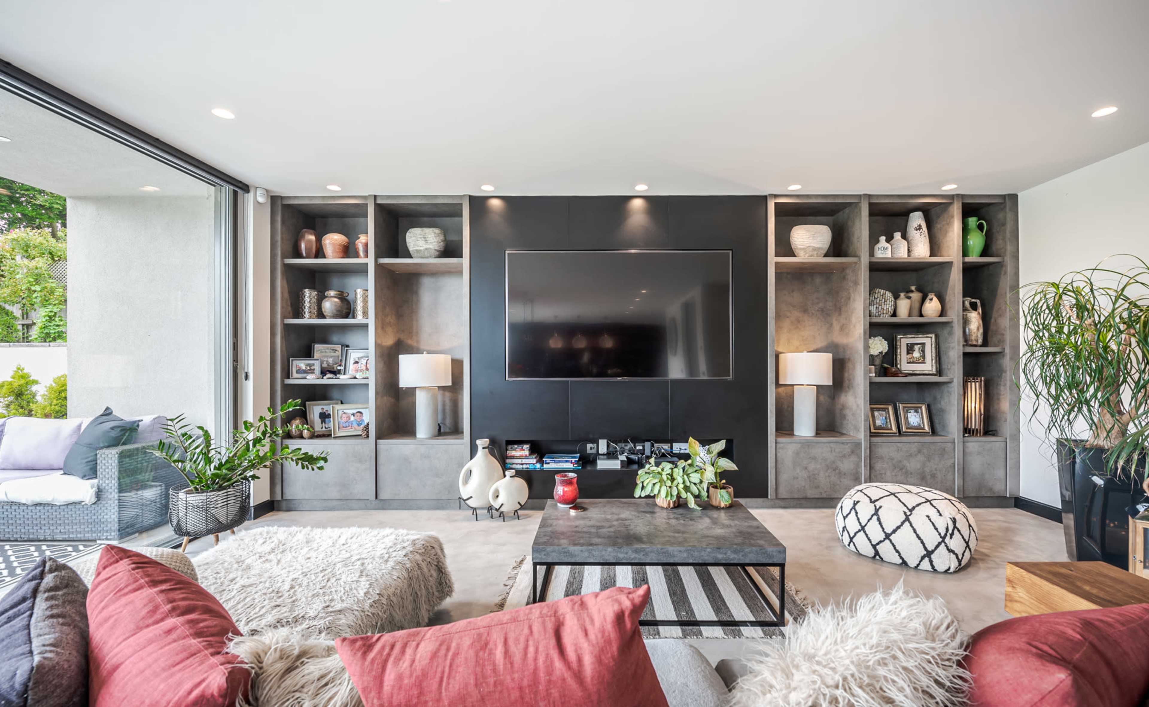 A modern living room features a large wall-mounted TV, built-in shelves with decorative items, and comfortable seating with colorful cushions.