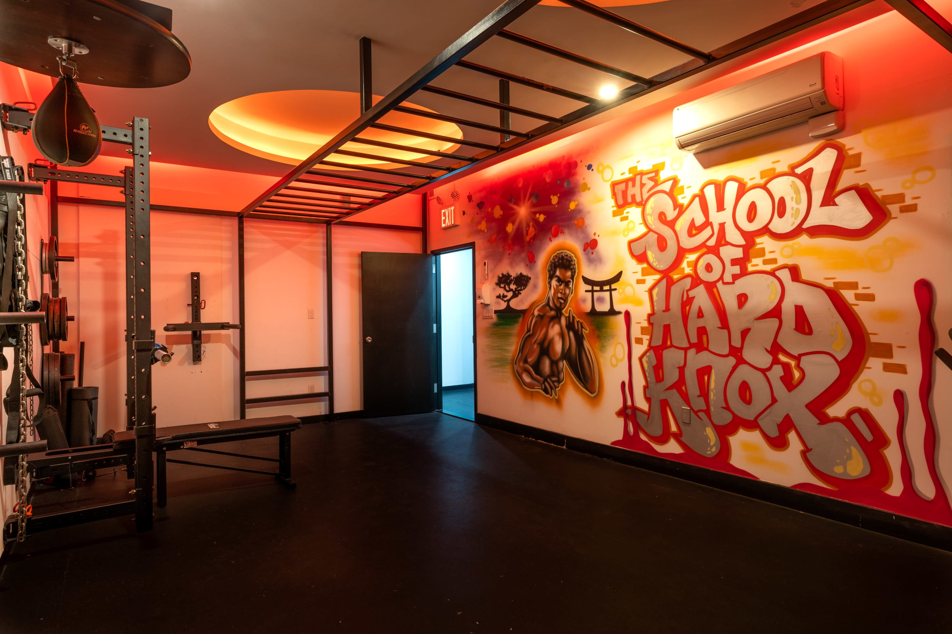 A gym interior featuring a wall mural that reads "THE SCHOOL OF HARD KNOCKS," along with punching bags and exercise equipment.