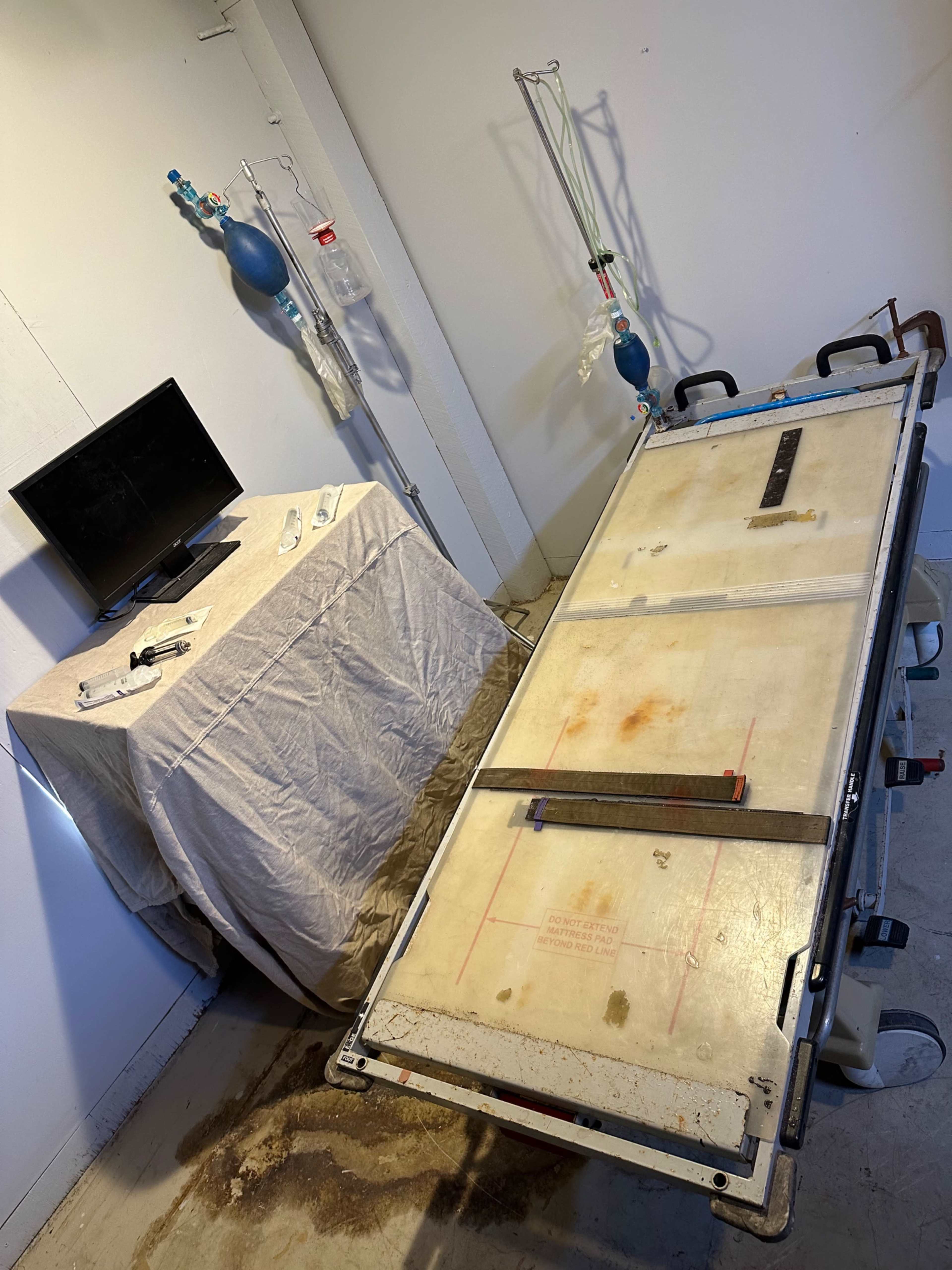A sterile examination room with an autopsy table, a draped table with medical equipment, and a monitor on the wall.