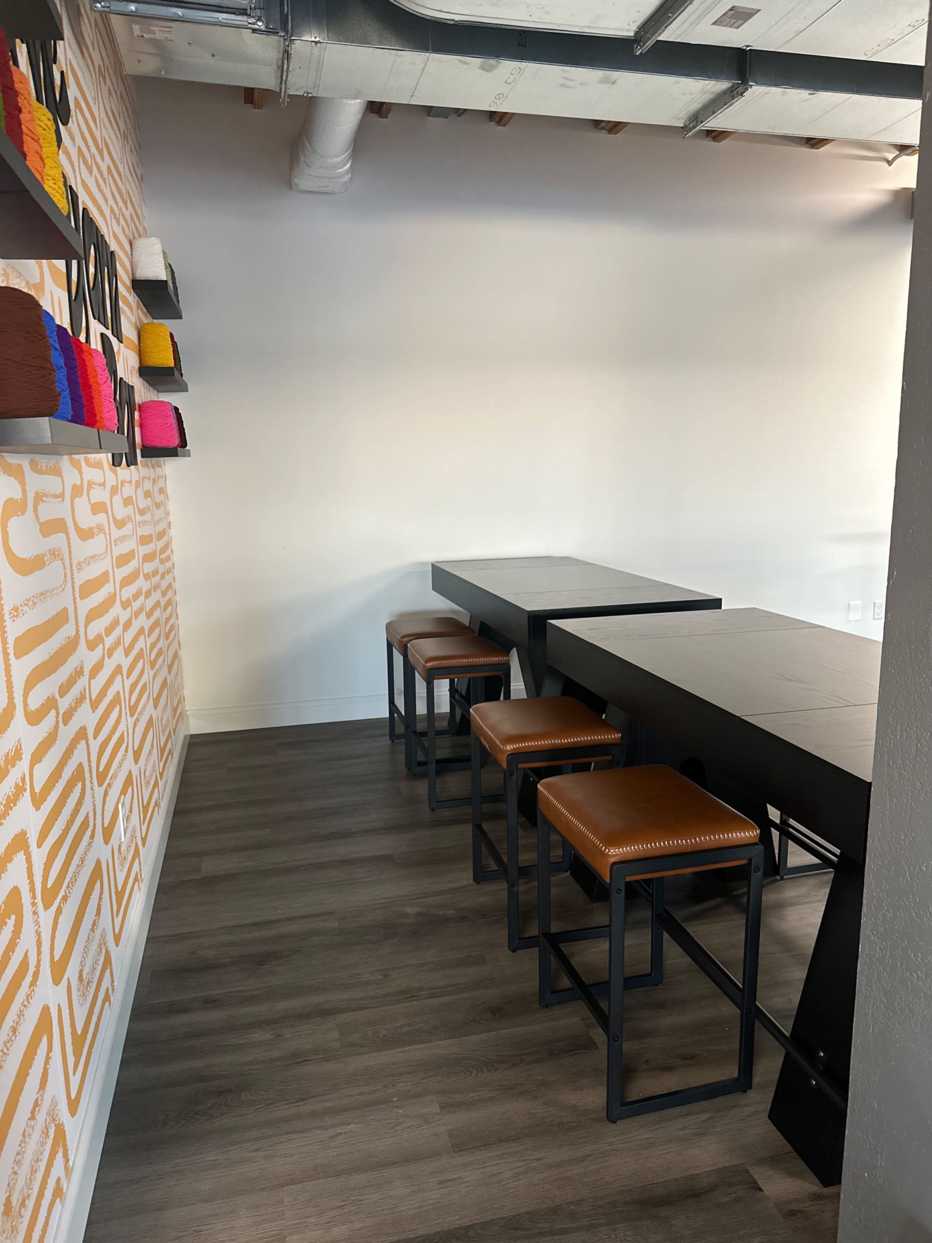 The image shows a modern interior space featuring a long black table with four brown stools against a wall adorned with colorful craft supplies.