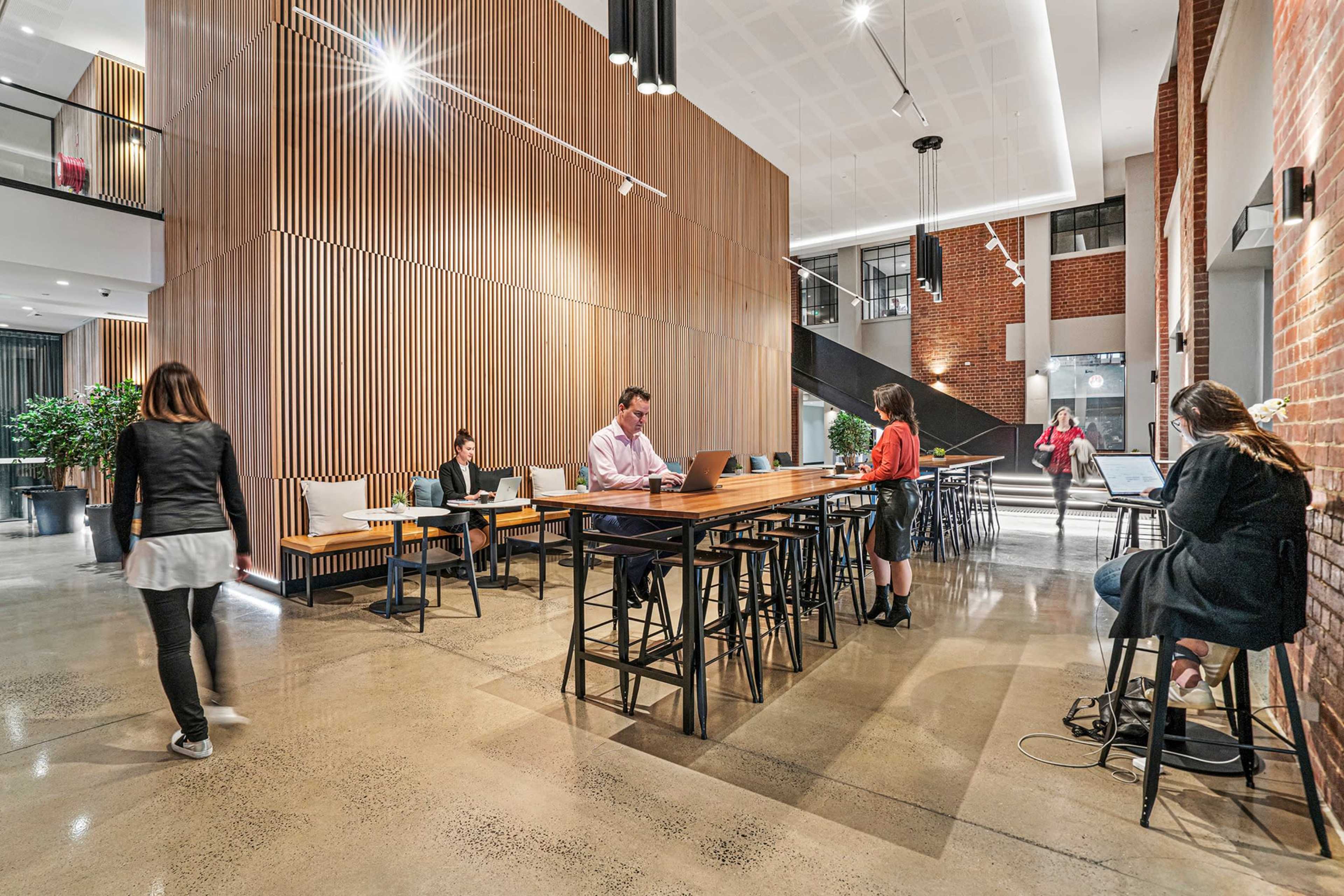 A modern workspace features wooden paneling and steel furniture, with individuals working on laptops at long tables and a staircase in the background.
