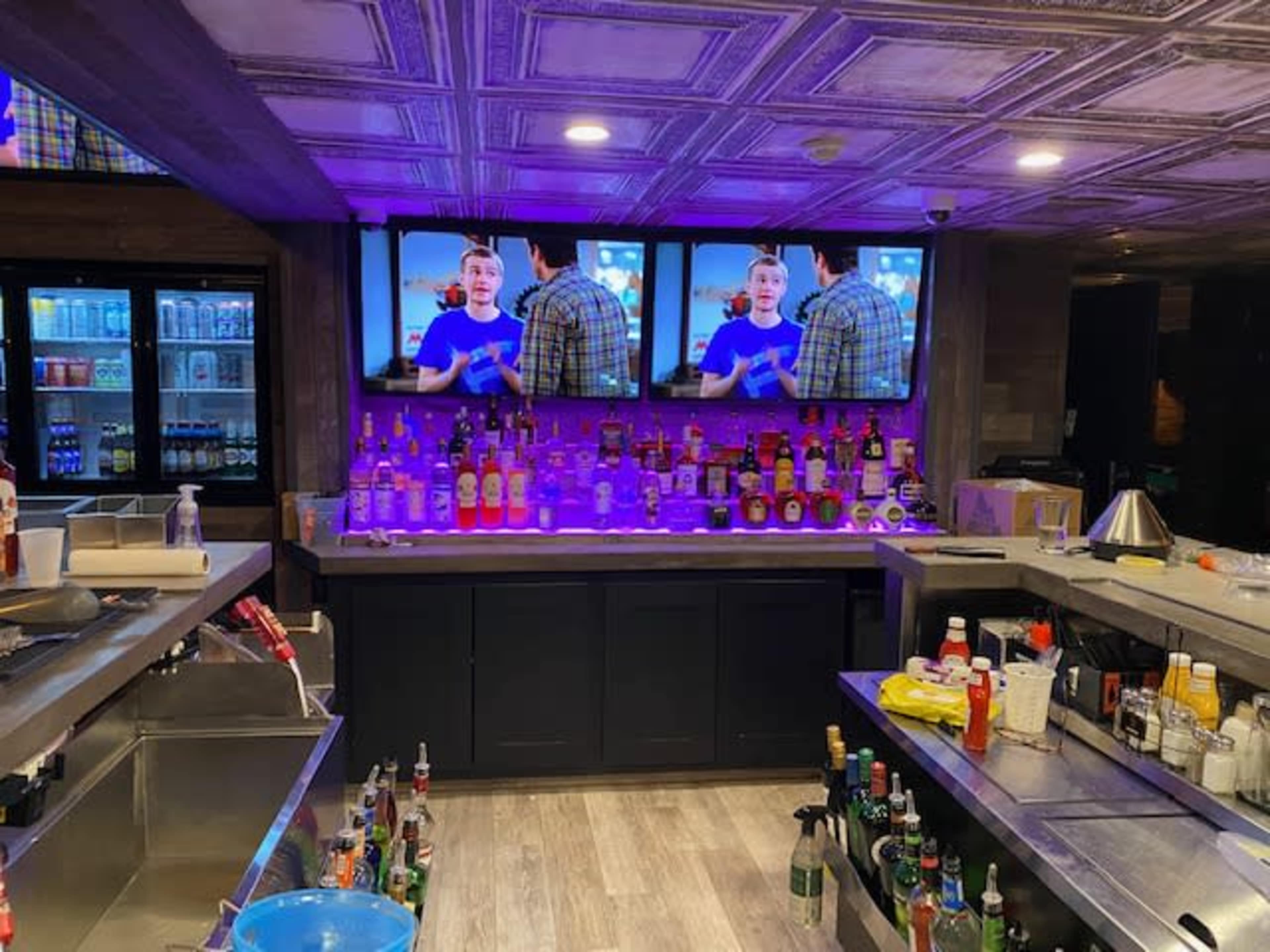 A bar area with a well-stocked liquor shelf, two televisions displaying a program, and various condiments and supplies on the counter.