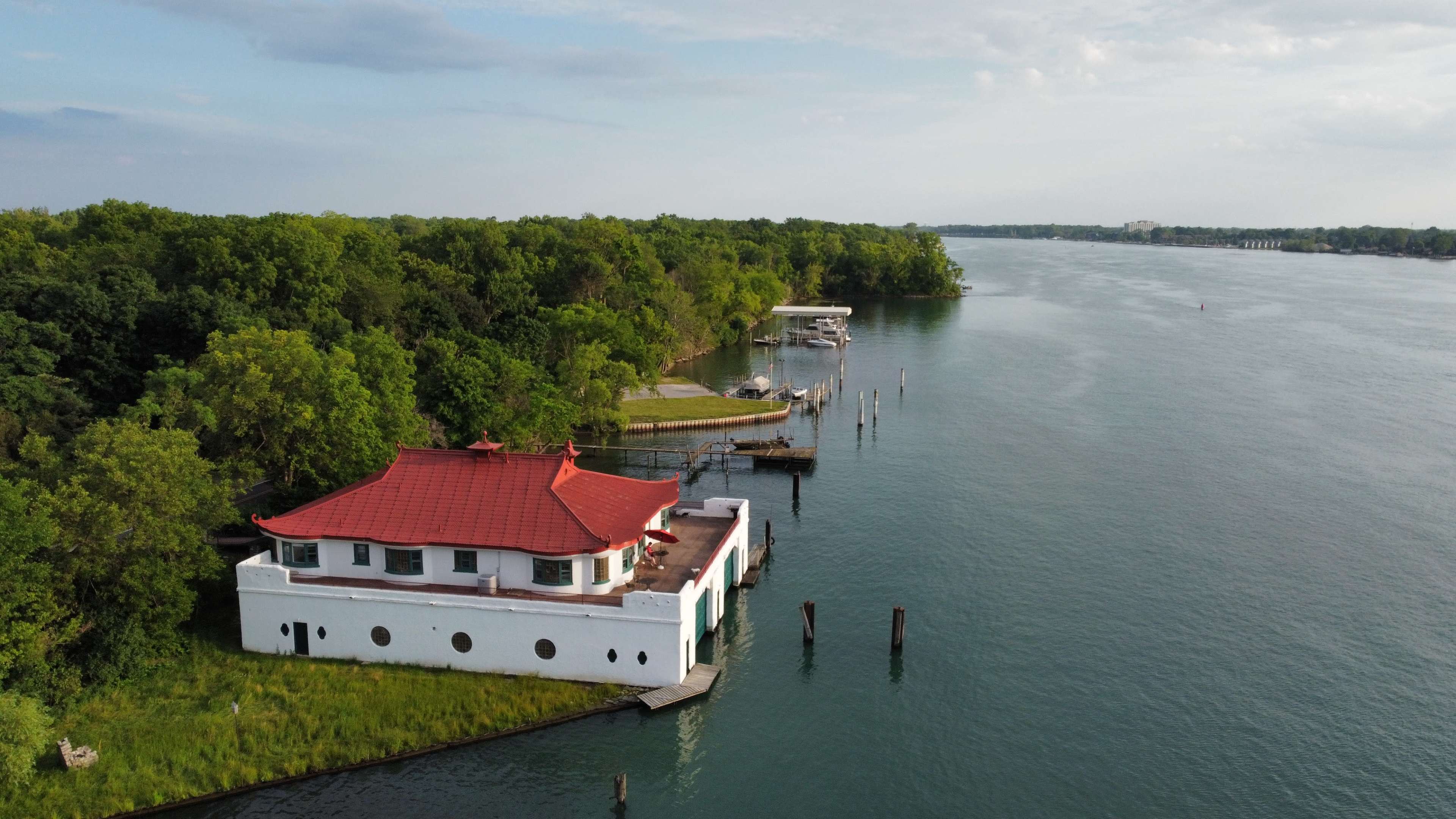 A white building with a red roof is situated on the bank of a river, surrounded by green trees and docked boats.