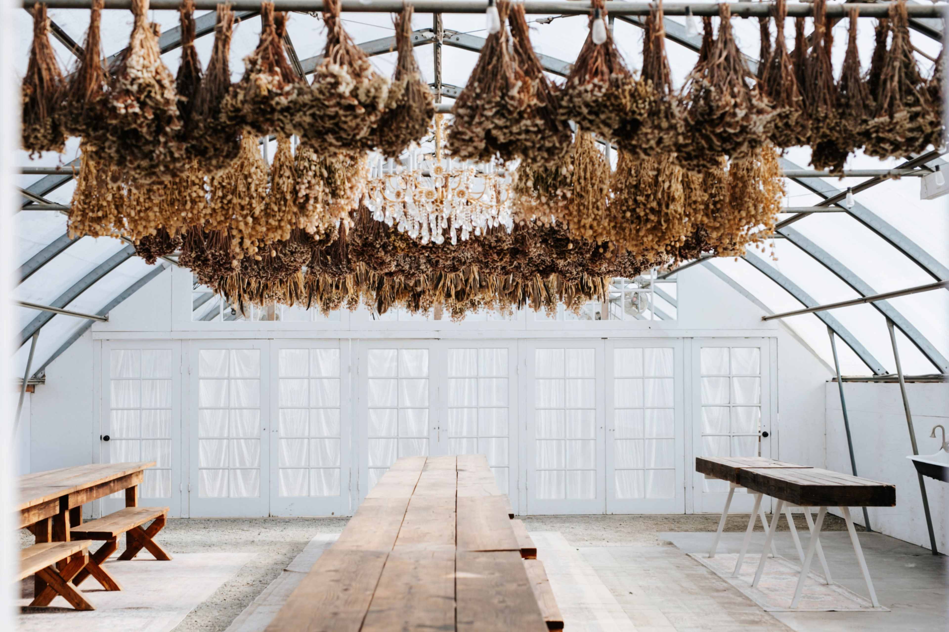 The image shows a spacious greenhouse with a wooden table and benches, adorned with hanging dried flowers from the ceiling.