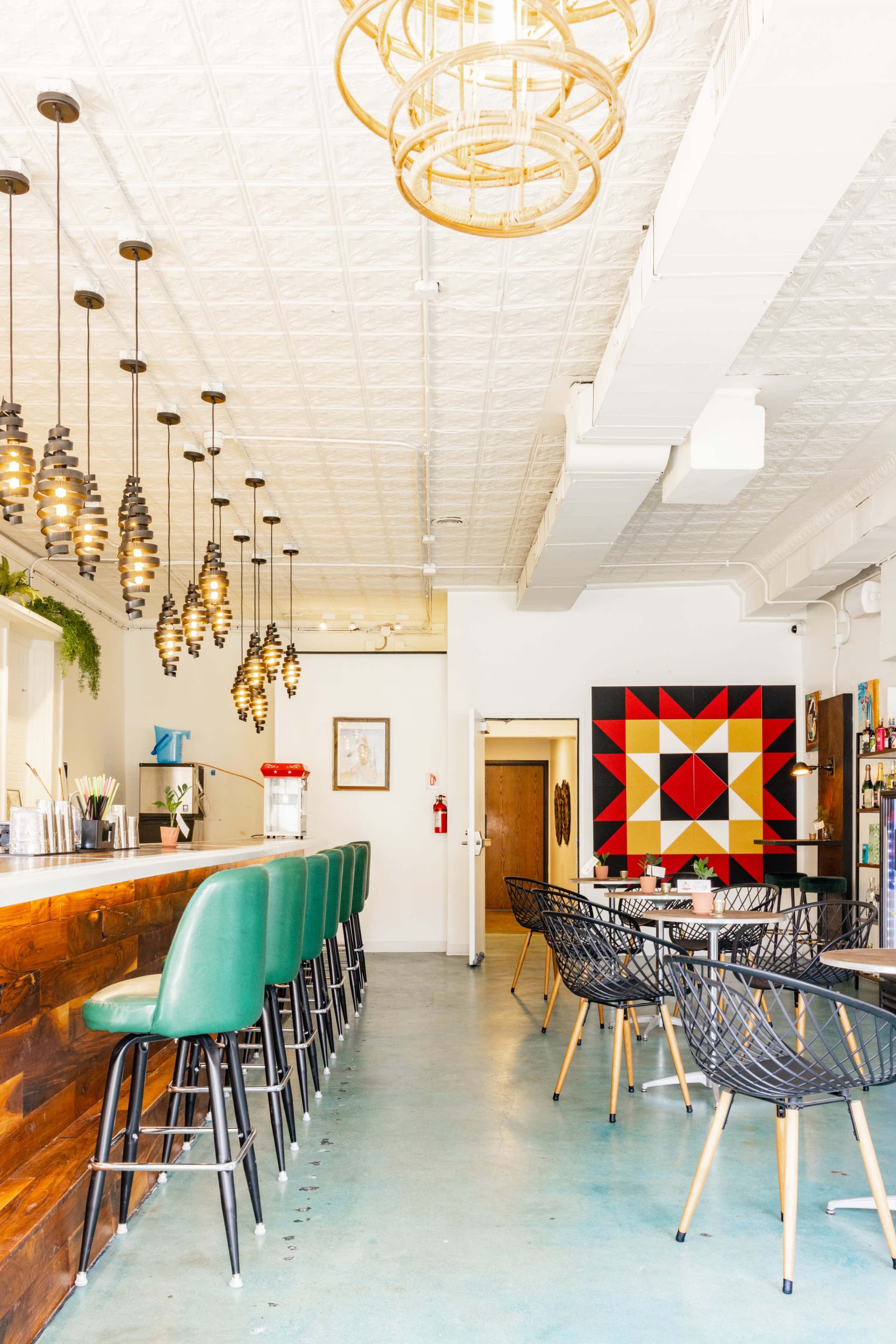 A modern café interior featuring a bar with green stools, stylish pendant lights, and a colorful geometric wall art.