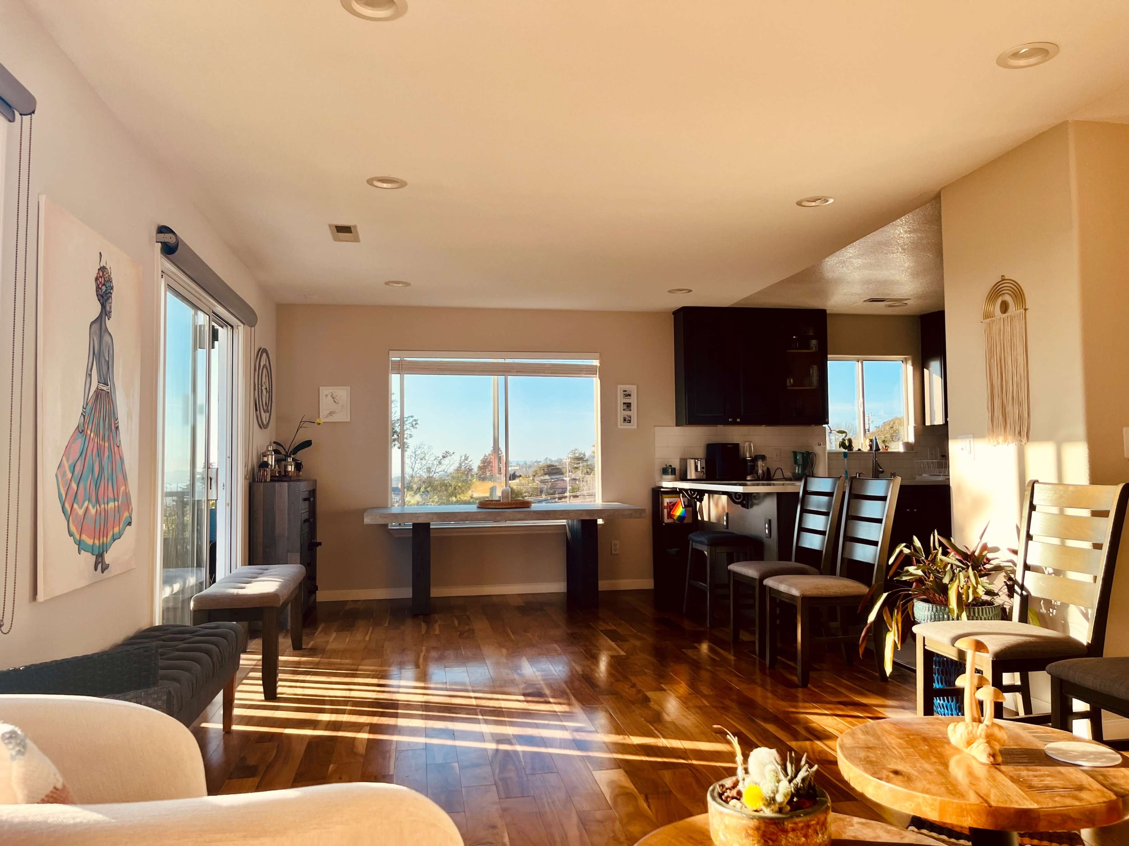 A bright living area features a table near a large window, surrounded by chairs and a kitchen bar, with wooden flooring and a colorful artwork on the wall.