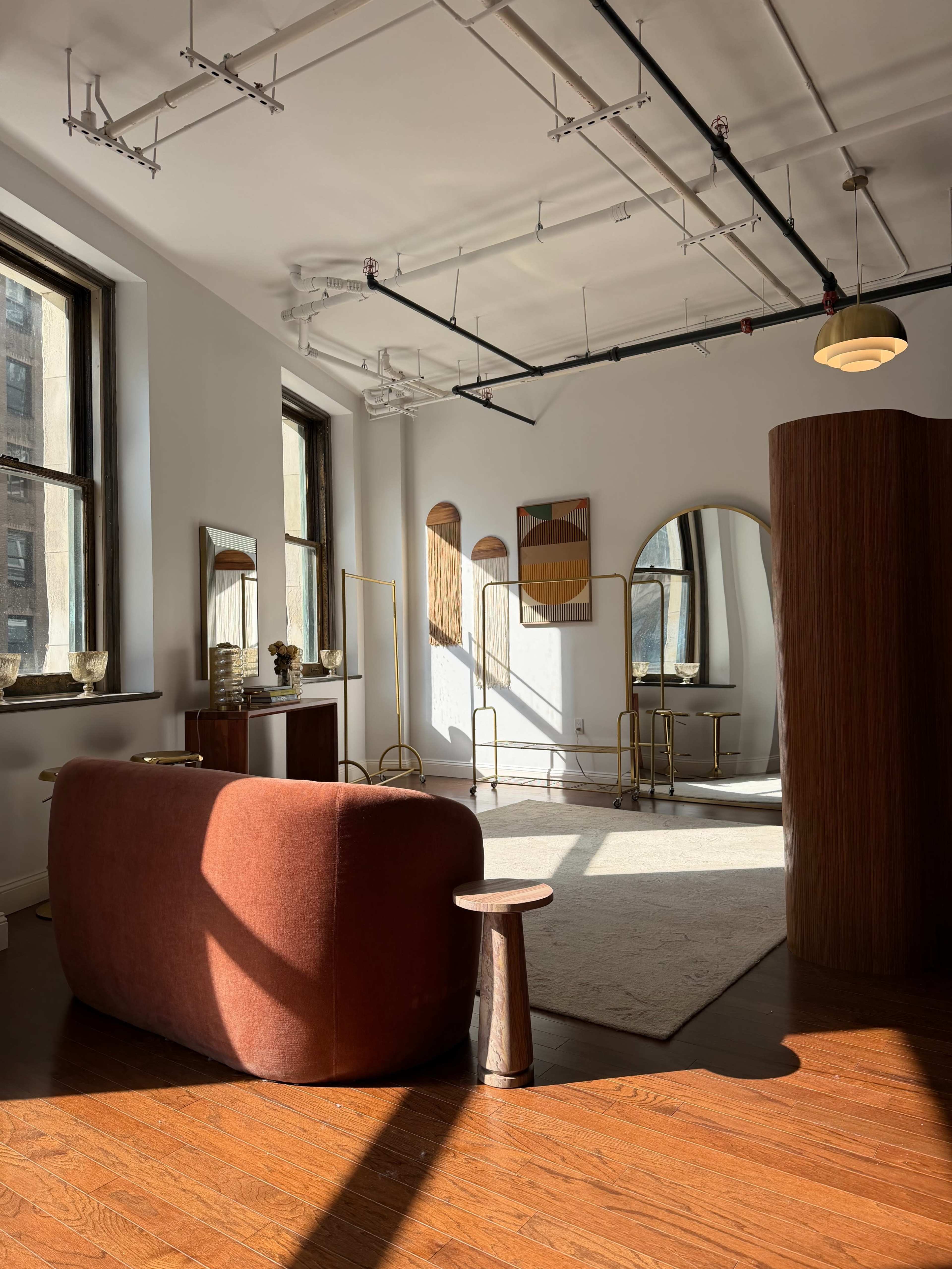 The image depicts a modern room with large windows, showcasing a reddish-brown sofa, a small wooden side table, and decorative mirrors on the walls.